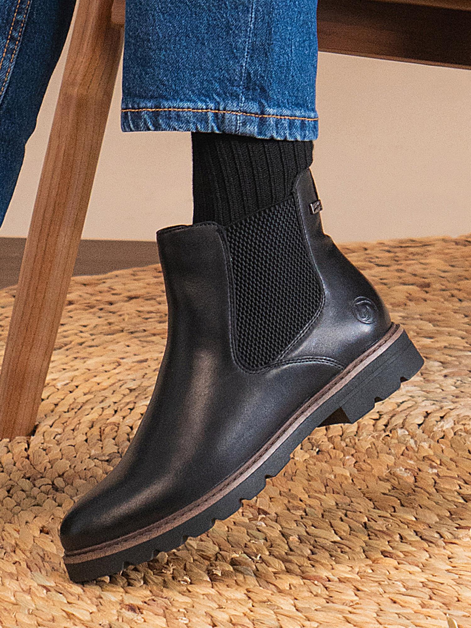 En person iført Remonte 04571 Torino Leather Ankle Boots, svarte sokker og blå jeans sitter på et vevd teppe med den ene foten på gulvet og det andre beinet lett bøyd.