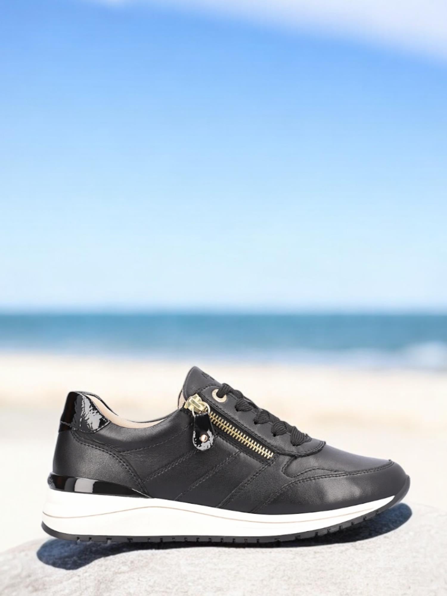 Remonte 06807 Noir Nappa-sneakerskoen, med svart skinn, en fleksibel hvit såle og gullfargede glidelåsdetaljer, er vist på en stein med en sandstrand og blått hav uskarpt i bakgrunnen under en klar himmel.