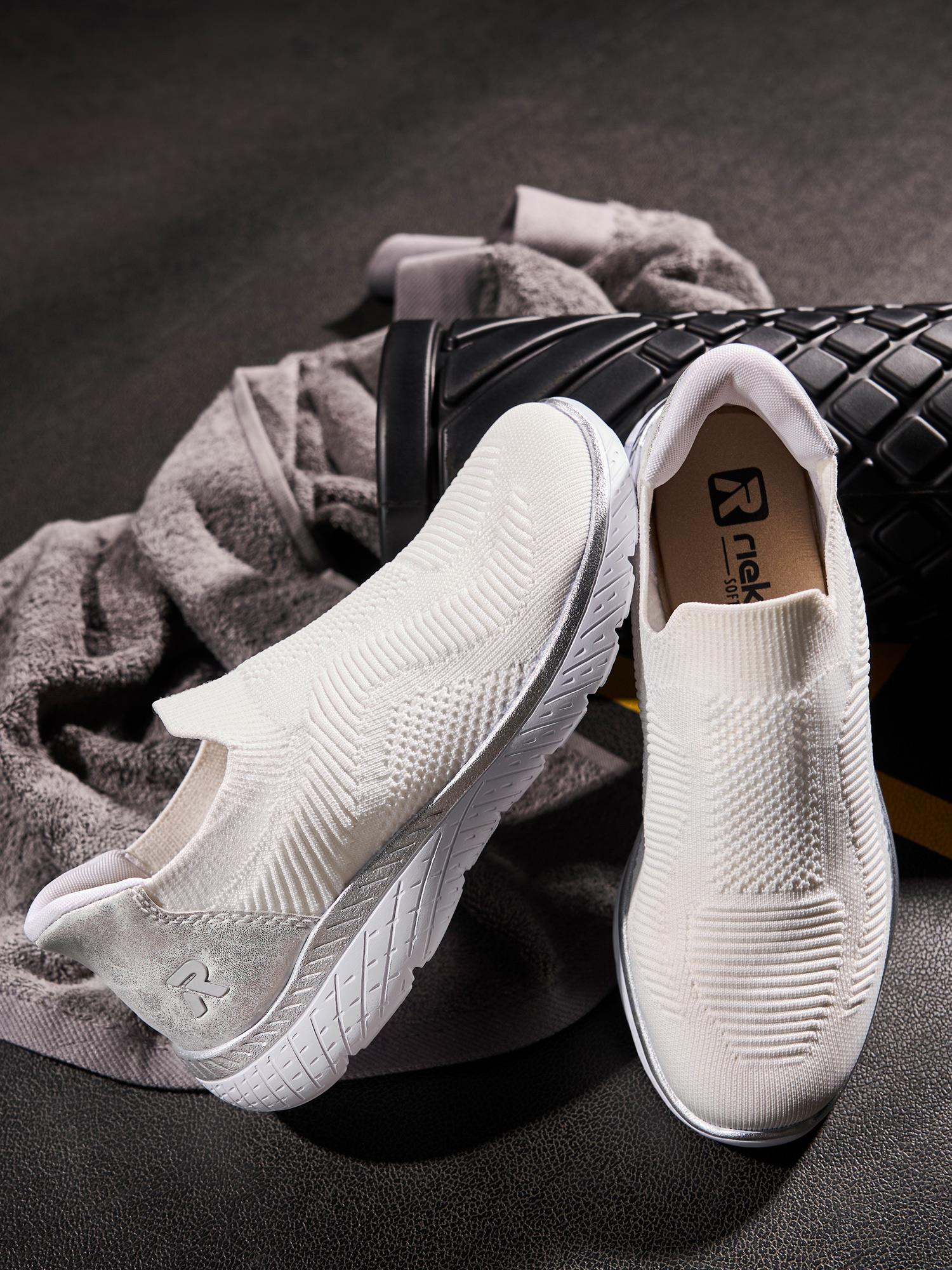 03238 Rieker Ready2Go Slip-on White joggesko fra Rieker Sport, med overdel i strukturstrikk, hviler på en mørk overflate ved siden av et grått håndkle og en svart vattert veske - perfekte pustende sko for din aktive livsstil.