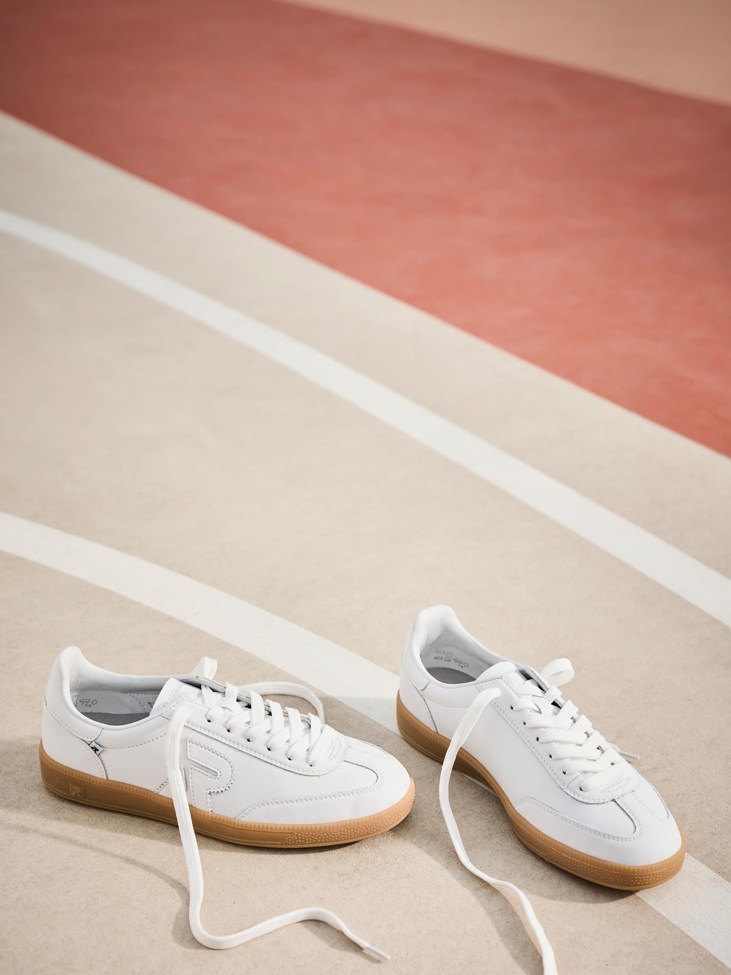 03486 Rieker San Diego Leather Sneaker Women White fra Rieker Sport har hvitt skinn, gummisåler og Swing2Go-teknologi. Sneakeren er vist på en banelignende overflate med buede hvite linjer, og har løse lisser for et avslappet utseende.