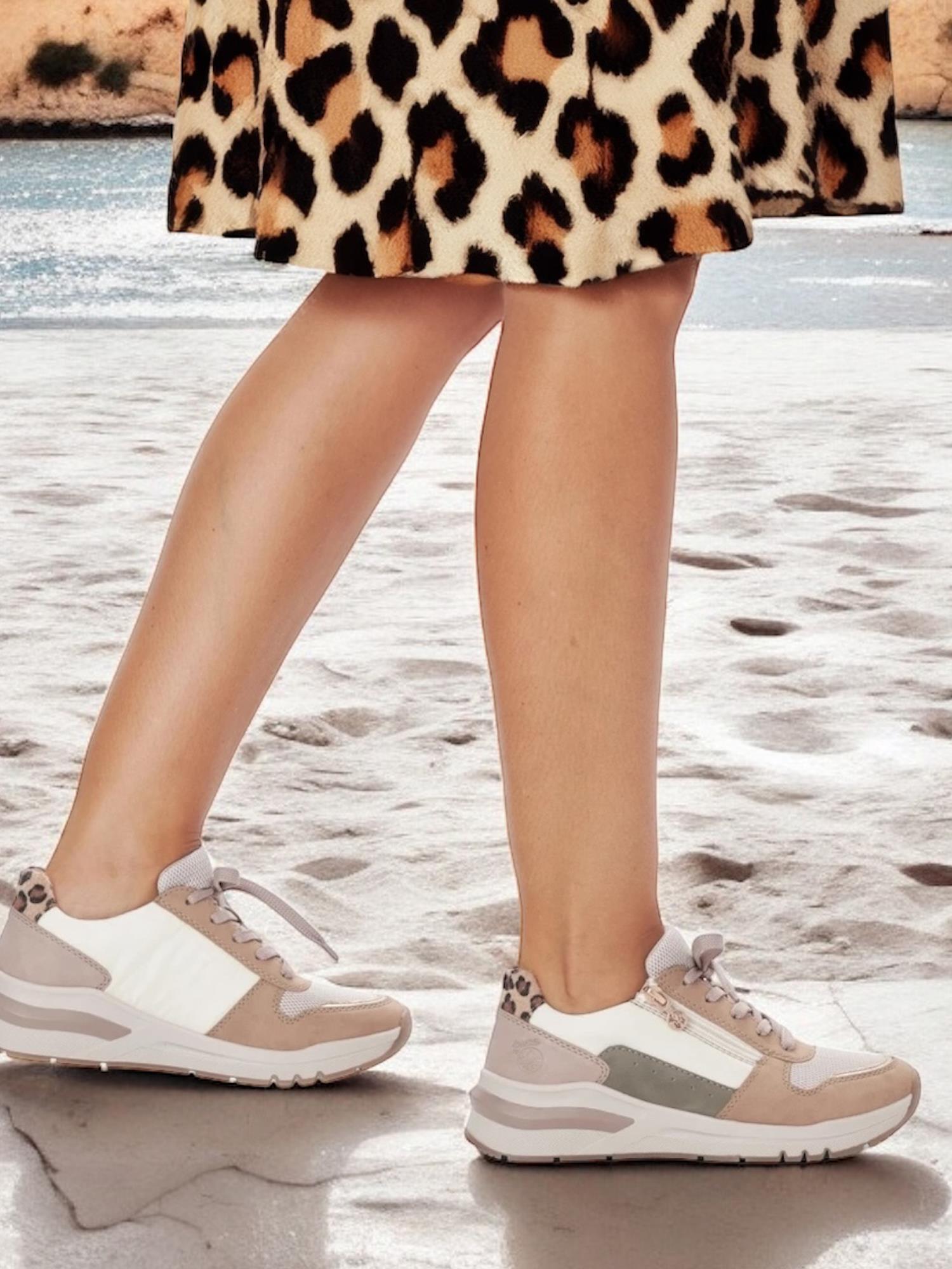 En person i et leopardprintskjørt og beige Rieker 06827 Sand Combo-sneakers med støtdempende såle fra Rieker Comfort & Style, med hvite og grå detaljer, går på sandgrunn nær vann. Bare bena og føttene deres er synlige.