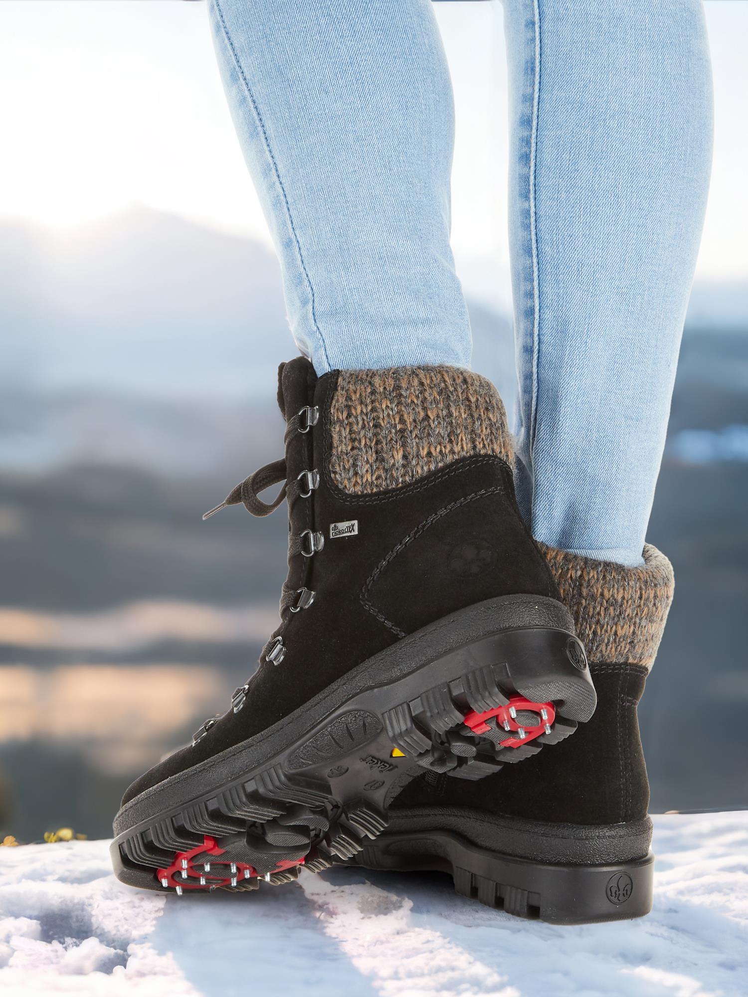 En person iført lyseblå jeans og 05364 Rieker Varmfòret vintersko med brodder fra Rieker Comfort & Style står på snø foran et uklart fjellandskap. Skoene har semsket skinn, varm fôring og tykk, gripesikker såle.