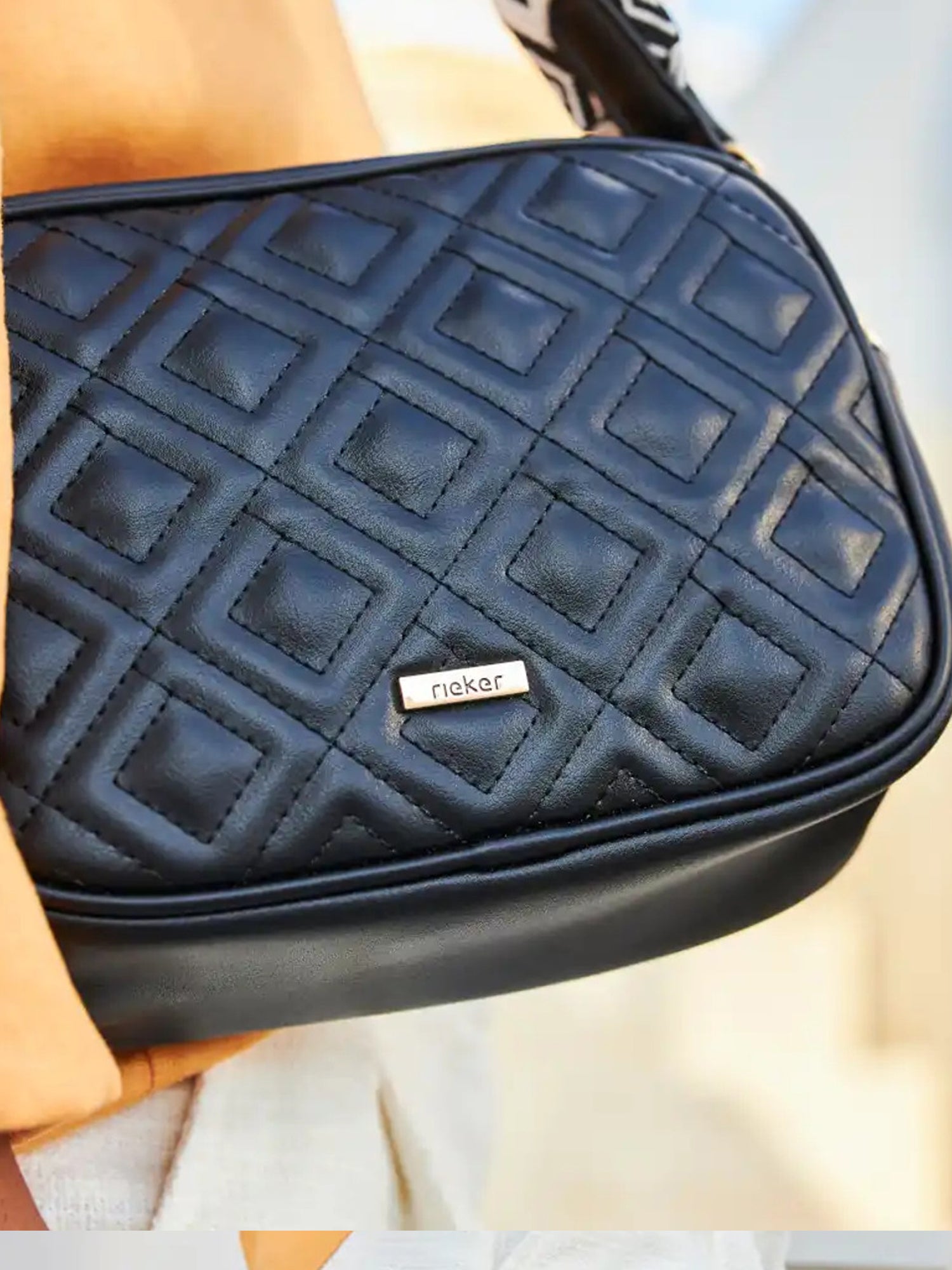 Et nærbilde av 02175 Rieker Melby Crossbody Mini Bag fra Rieker Comfort & Style, en svart veske med diamantquiltet mønster og metallmerke, stylet med et solbrunt antrekk.