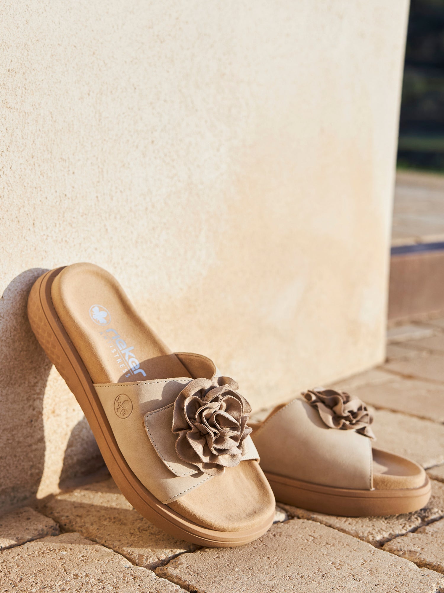 02193 Rieker Sandelle Sandal fra Rieker Comfort & Style, i beige med dekorative stoffblomster og en lett TR-sålen for sommerkomfort, hviler på en steinterrasse mot en lys vegg i solen.