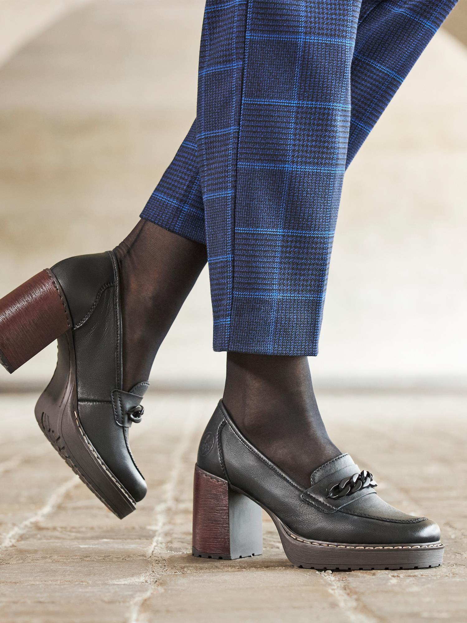 En person står på et flislagt gulv iført mørke, gjennomsiktige strømpebukser, blårutede bukser og Rieker Comfort & Style 04394 Montpellier Loafer Pumps.