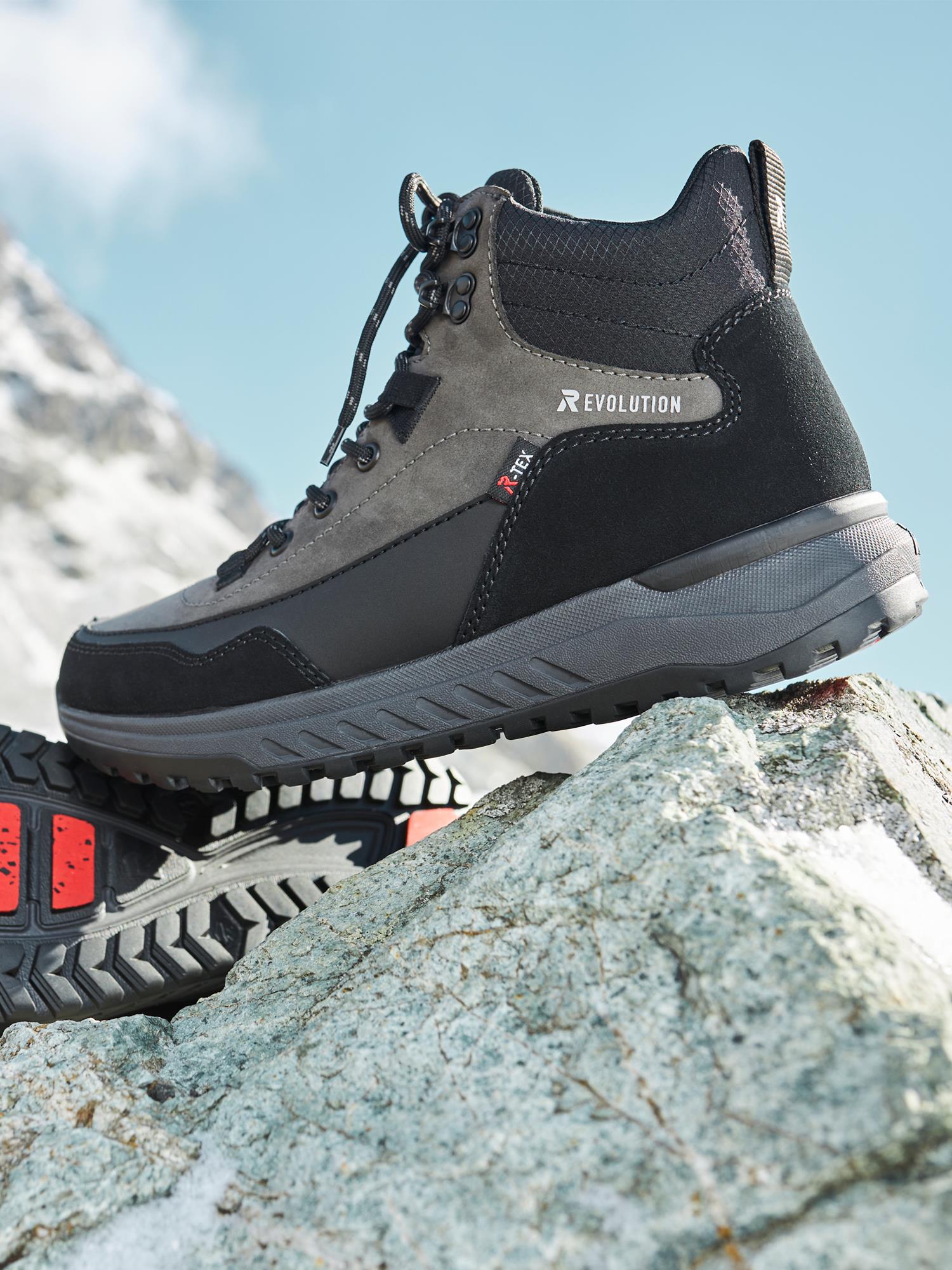 04505 Rieker-Evolution Coal Boot Tex-Membran Ice-Grip fra Rieker Sport er vist utendørs på steiner, der den robuste sålen og det værbestandige designet fremheves mot et bakteppe av fjell og blå himmel.