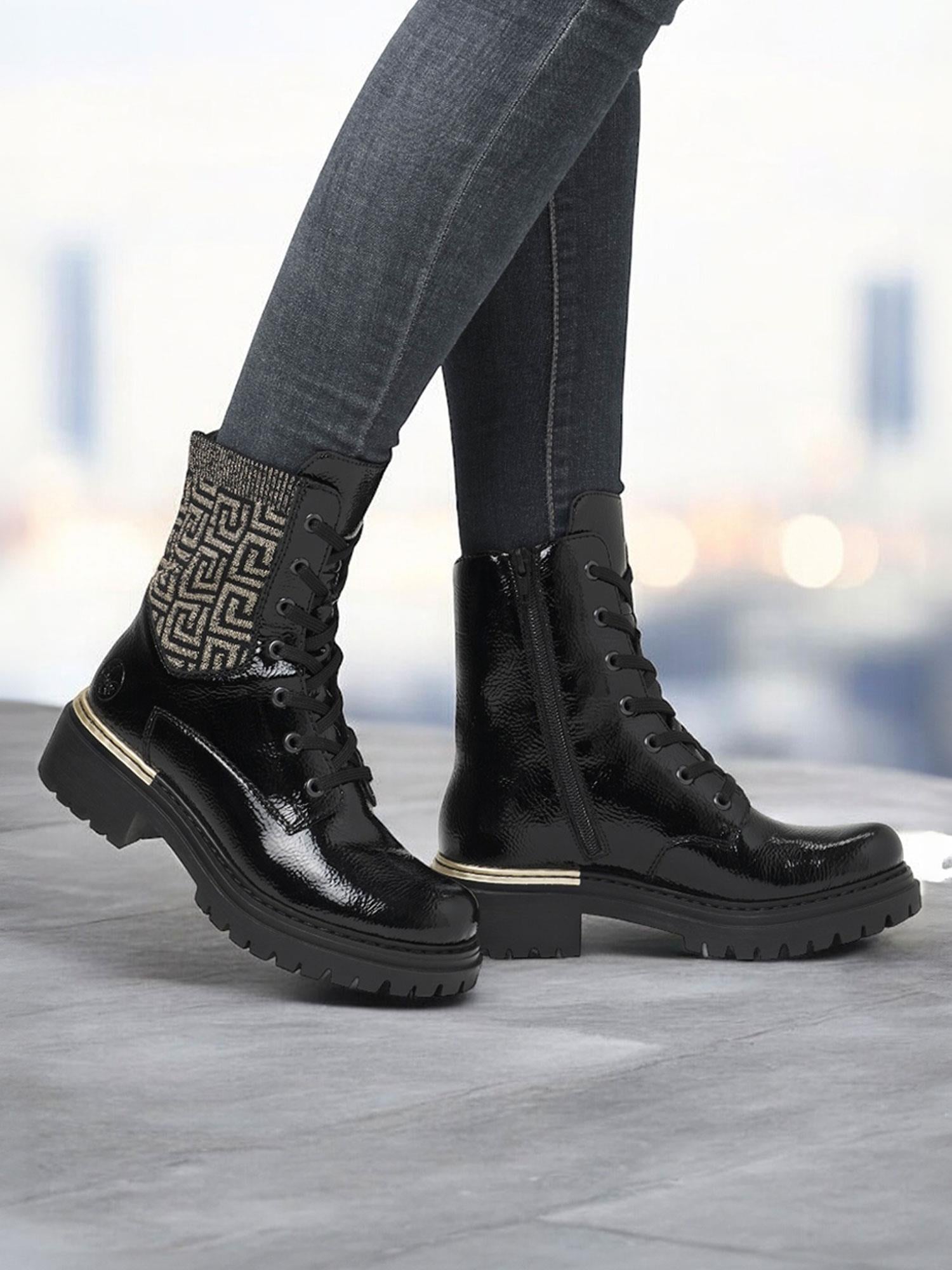 En person iført mørke skinny jeans og Rieker Comfort & Style 04740 Rieker Artemis Zip Boots med tykke såler og mønstret stoffdetalj nær toppen, står på et grått underlag mot en uskarp bybakgrunn.