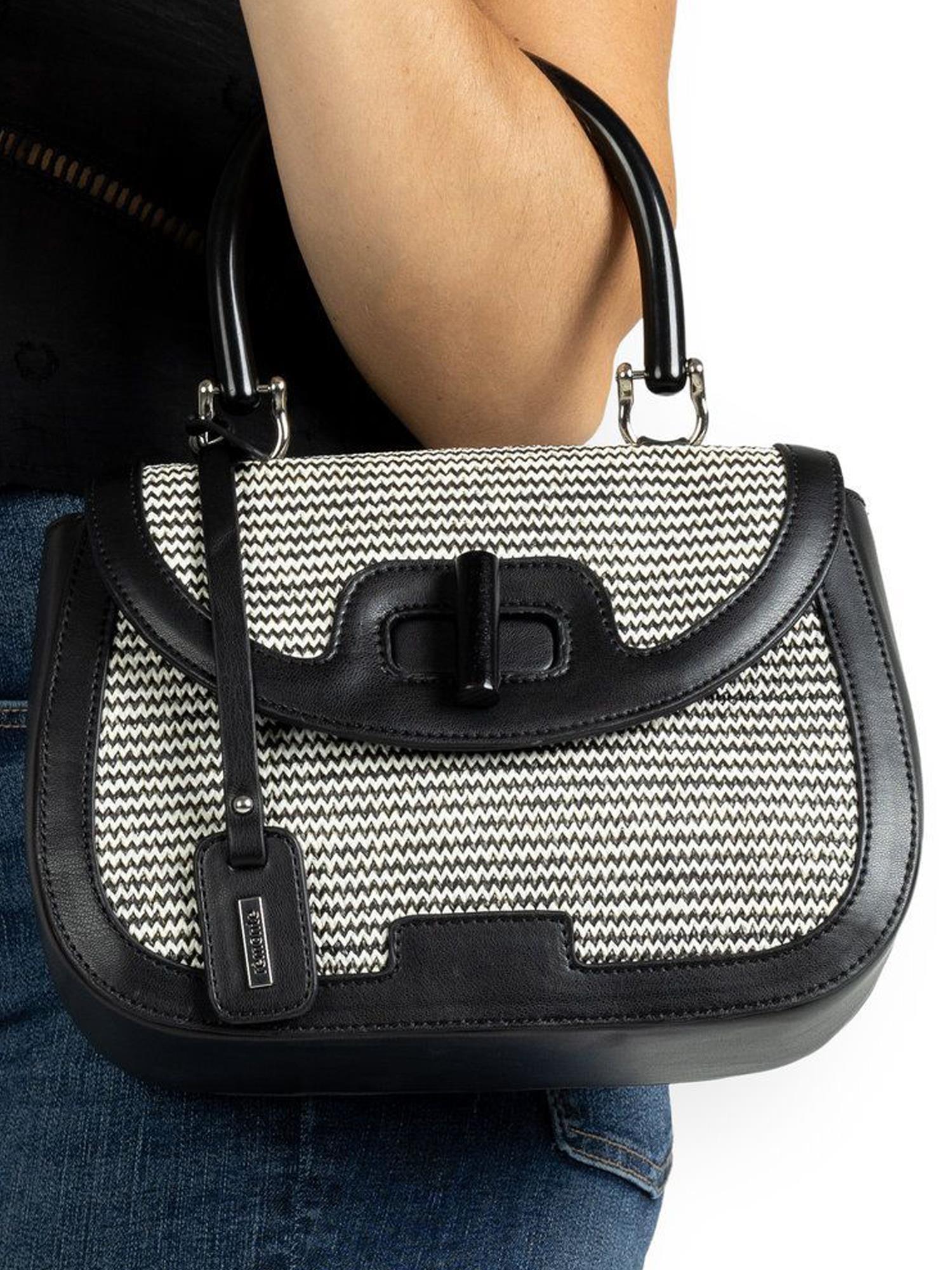 En person holder en 03715 Rieker Siradia Handbag Svart by Remonte, med svart og hvitt vevd design, svart kant og håndtak, dekorativ spenne og hengelapp. Vedkommende har på seg blå jeans og en svart topp.