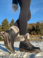 En person i svarte leggings og Remonte 04794 vintersko skinn ullfôret Fiber-Grip Sort står på snø, med trær og en bakke bak seg under en klarblå himmel.