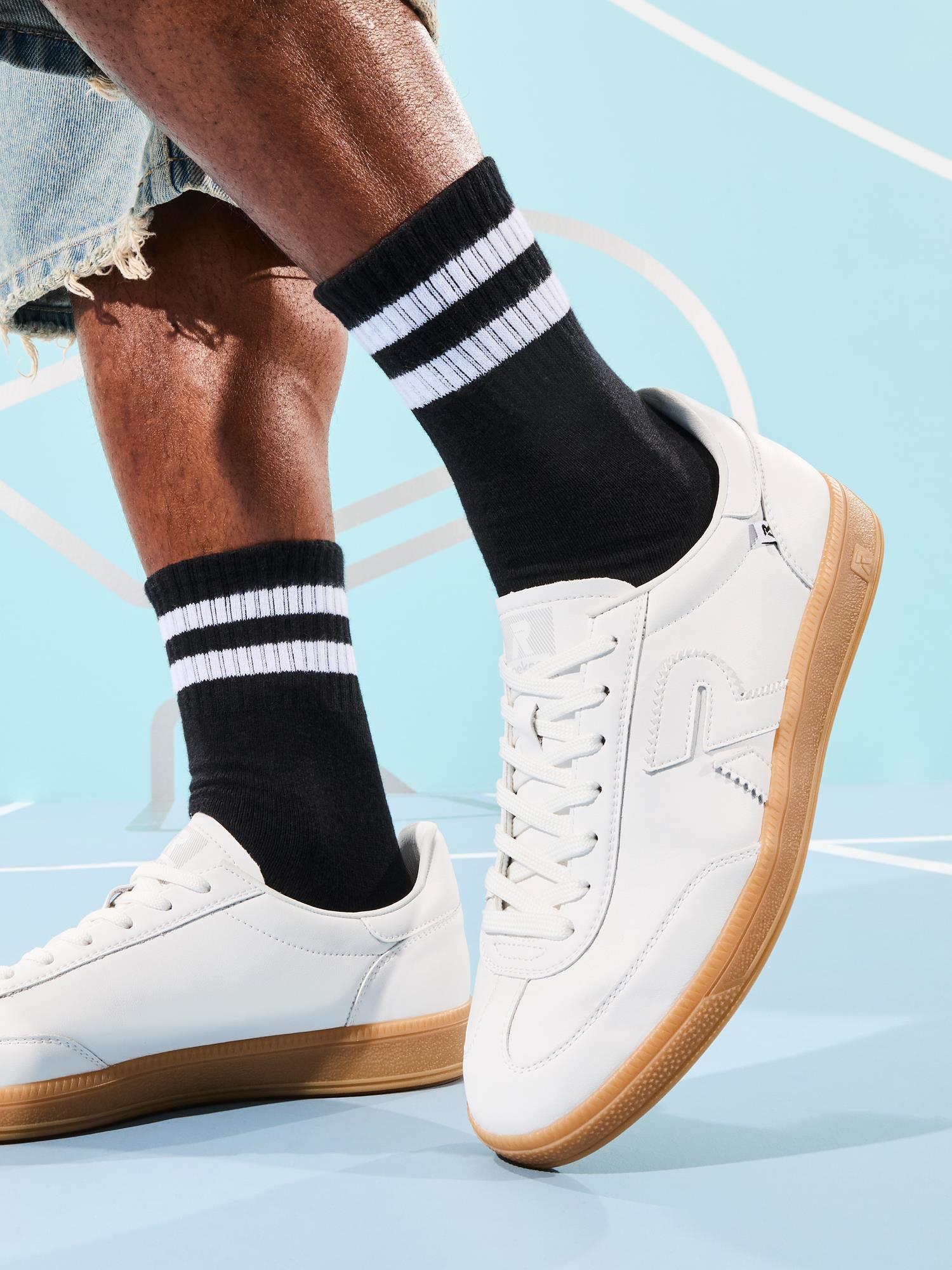 En person viser en uformell stil iført Rieker 03336 Rieker San Diego Leather Sneaker Herre Hvit med gummisåler og svarte sokker, sammen med frynsete lyseblå denimshorts mens han står på et lyseblått underlag.