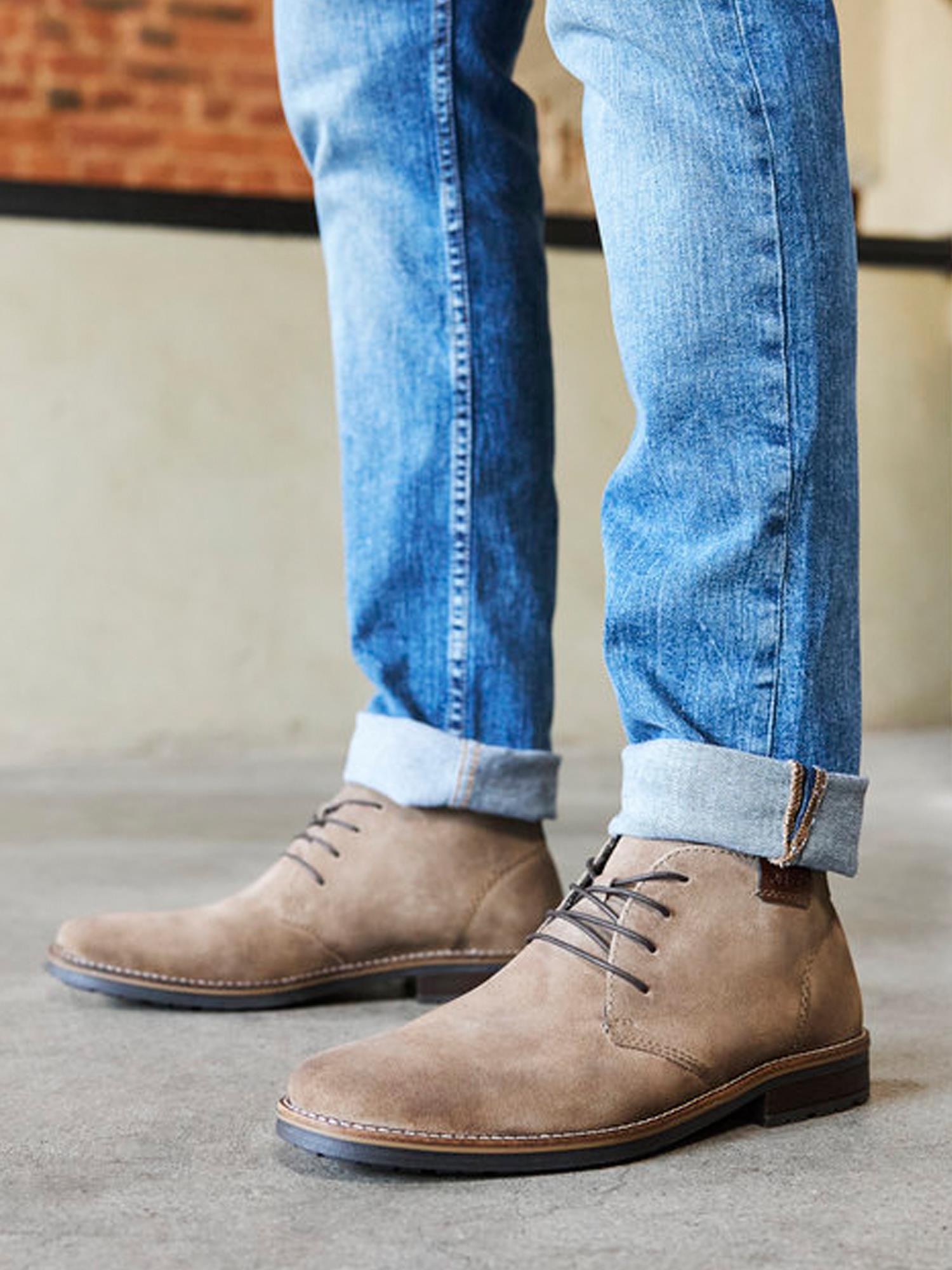En person i Rieker Comfort & Style 04494 Mens Suede Short Boots og blå jeans med mansjetter står innendørs på betong, med en uskarp murvegg bak – noe som representerer moderne komfort.