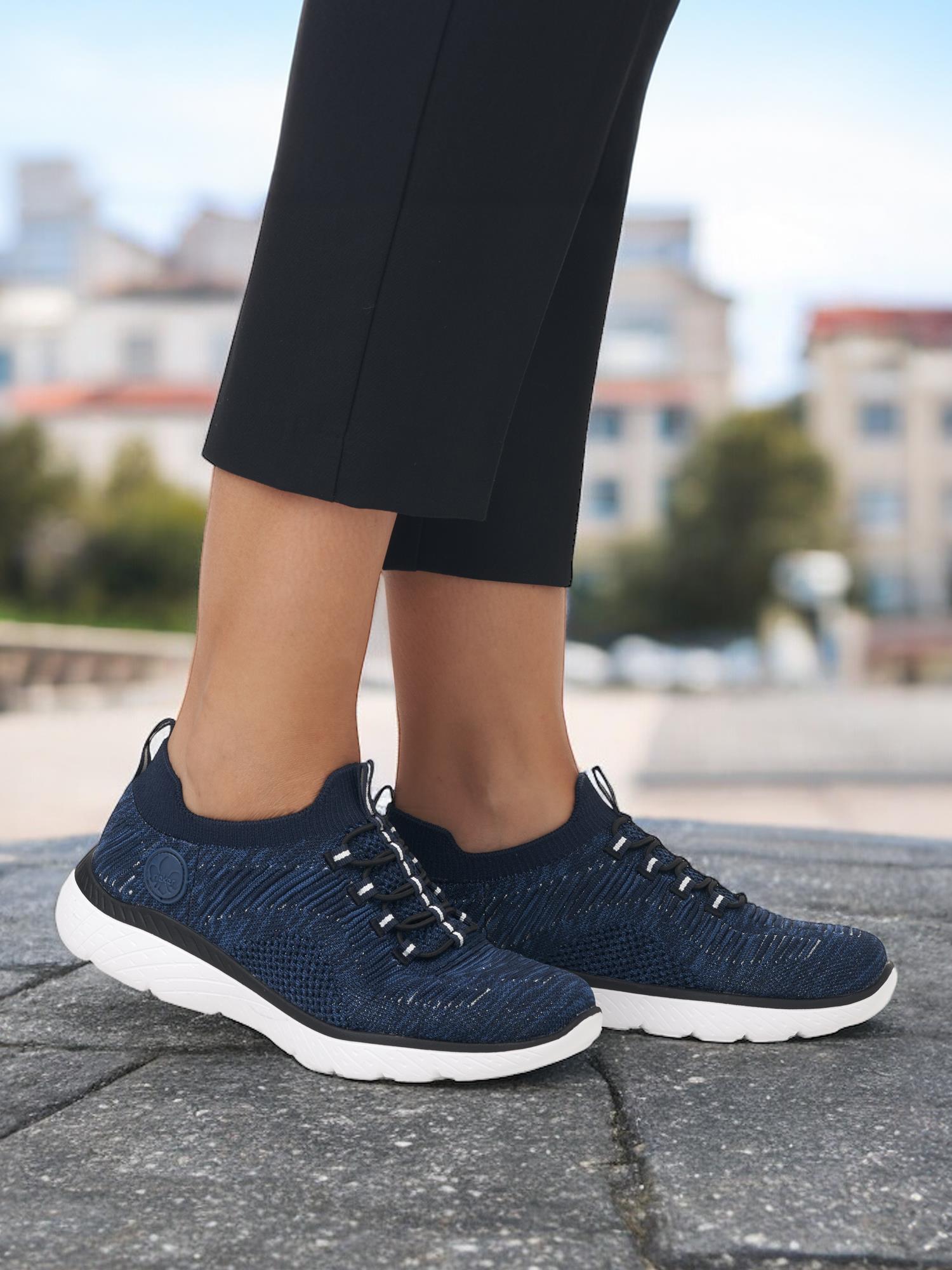 En person i mørkeblå 07488 Rieker Breathe Sneakers fra Rieker Comfort & Style, med hvite såler og svarte bukser, står utendørs på steinbelegg. Sneakersene har Antistress-teknologi og MemoSoft-innersåle for komfort.