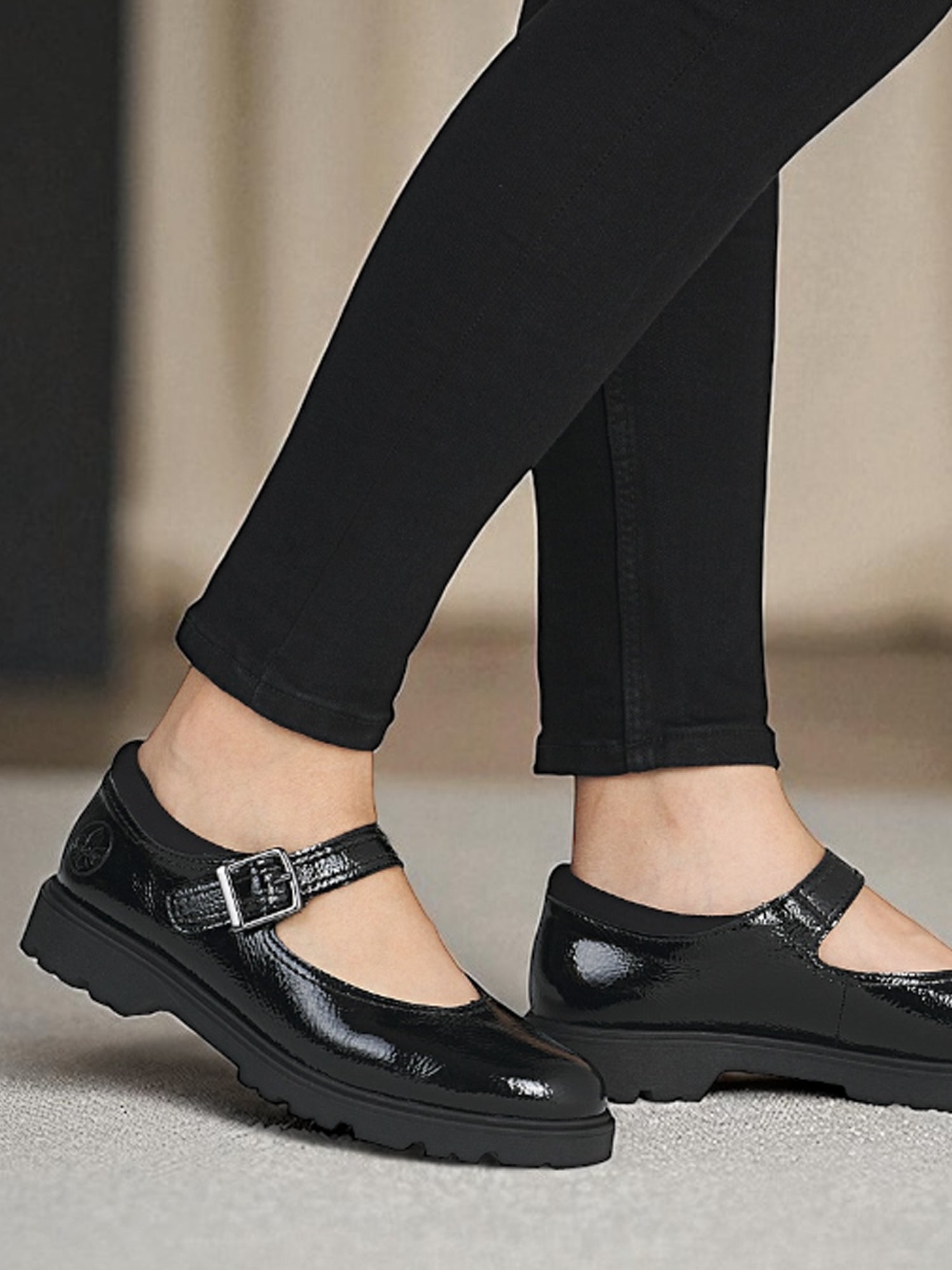 Noen i svart, skinnende 04289 Rieker Mary Jane Sko fra Rieker Comfort & Style, med tykke såler og sølvspenner, sammen med svarte leggings, står på et lyst teppe.