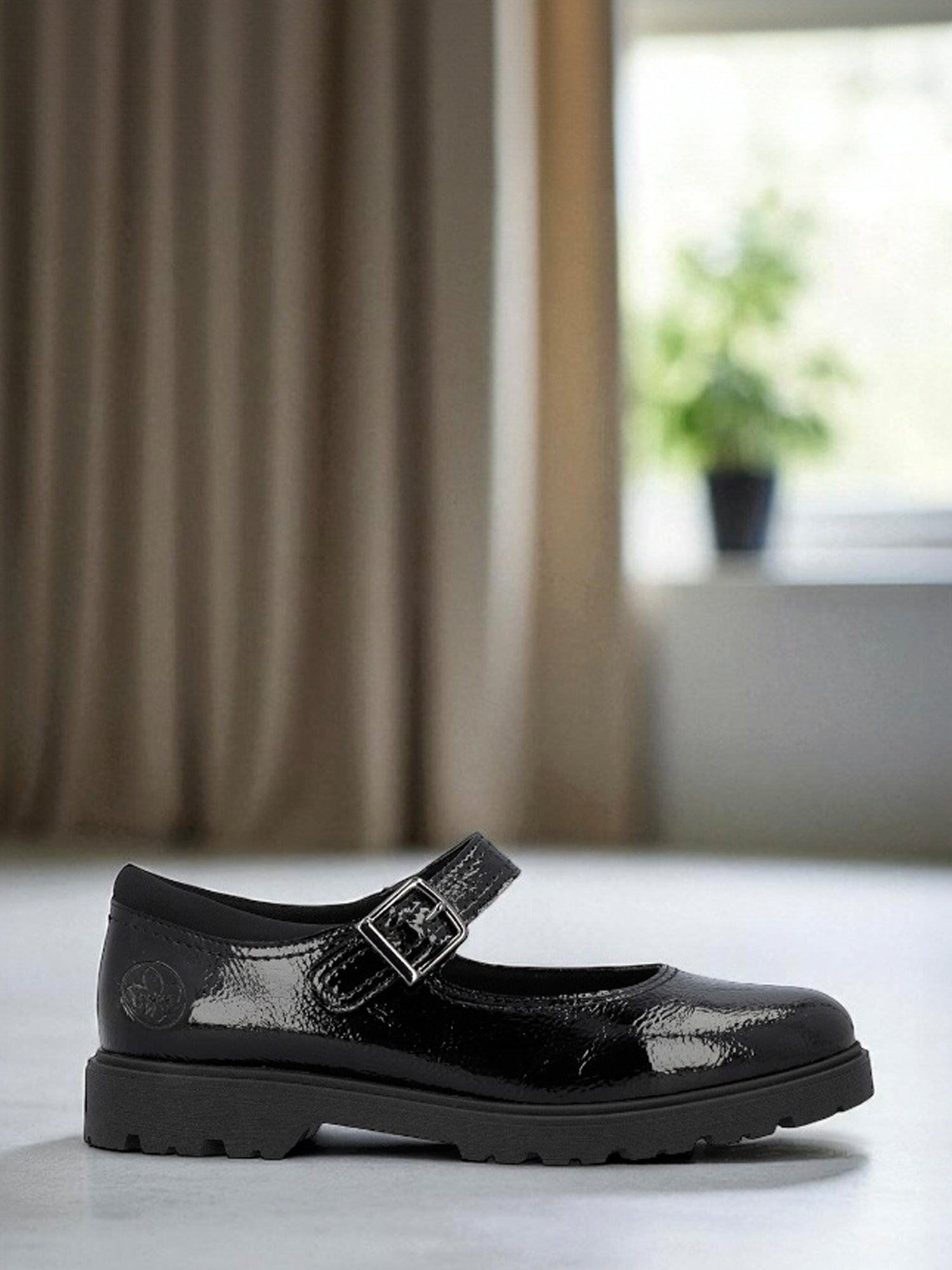 04289 Rieker Mary Jane Sko fra Rieker Comfort & Style har en skinnende svart finish, tykk såle og spennerem. Denne skoen er vist innendørs på en lys flate med beige gardiner og en potteplante, og utstråler både stil og komfort sko appeal.