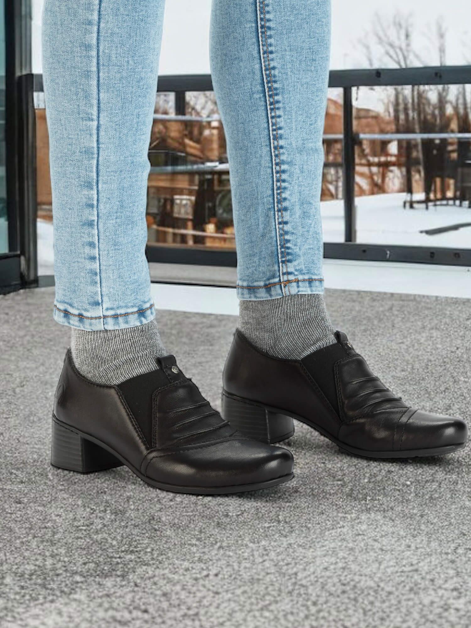 En person iført lyseblå jeans, grå sokker og svarte 04372 Rieker Skinn Mokkasiner fra Rieker Comfort & Style med spesiell skinninnersåle og antistressteknologi, står på et grått teppe foran en snødekt utendørsscene som er synlig gjennom glass.