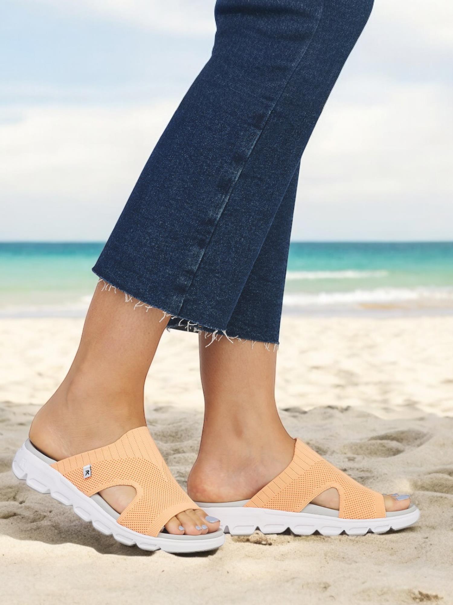 En person i avkortede blå jeans har på seg Rieker-Evolution 07524 Sunflare Sandal Oransje, og står på en sandstrand med havet og himmelen i bakgrunnen.