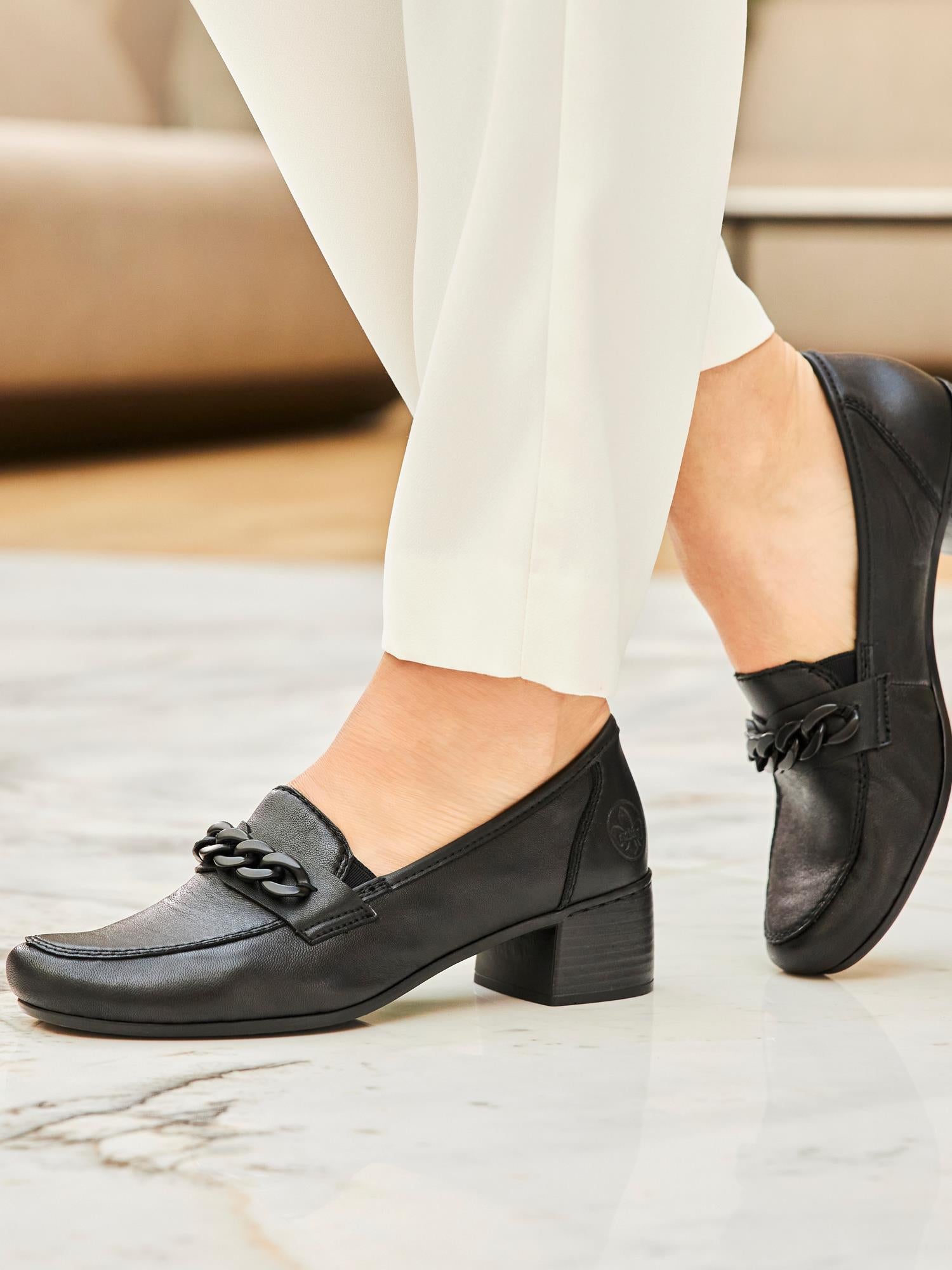 En person iført hvite bukser og svarte 07640 Rieker Midnight Leather Loafers fra Rieker Comfort & Style, med en kraftig kjededetalj og polstret såle, står på et polert marmorgulv.