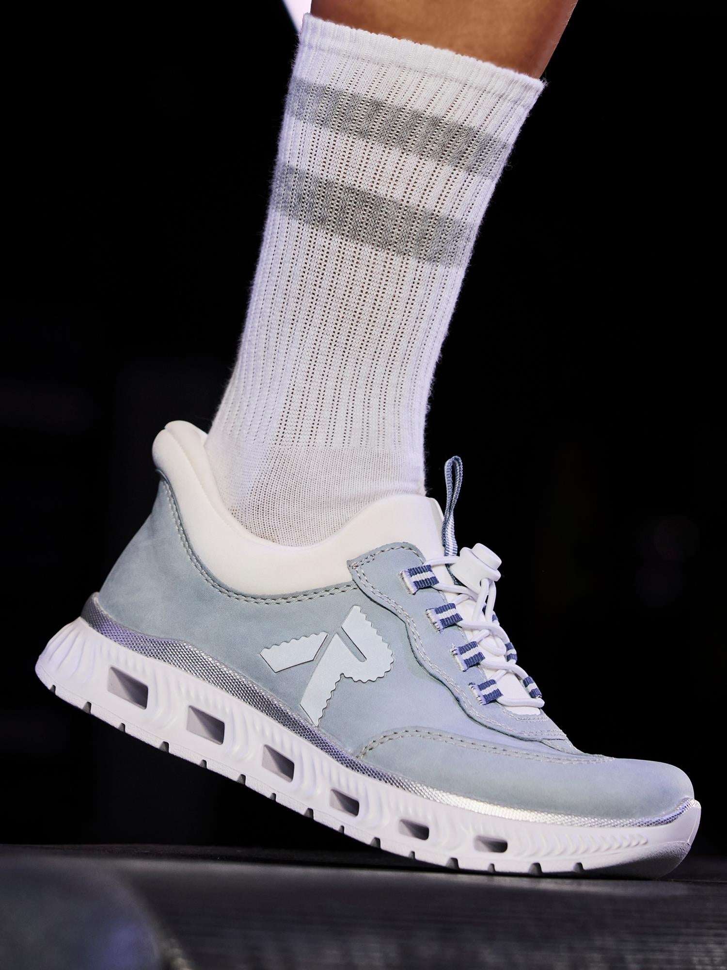 Et nærbilde viser en person iført lyseblå 03278 Rieker Ready2GO Slip-On Sneakers fra Rieker Sport, med hvite lisser, en tykk hvit såle og den karakteristiske R-logoen, stylet med hvite sokker med to grå striper.