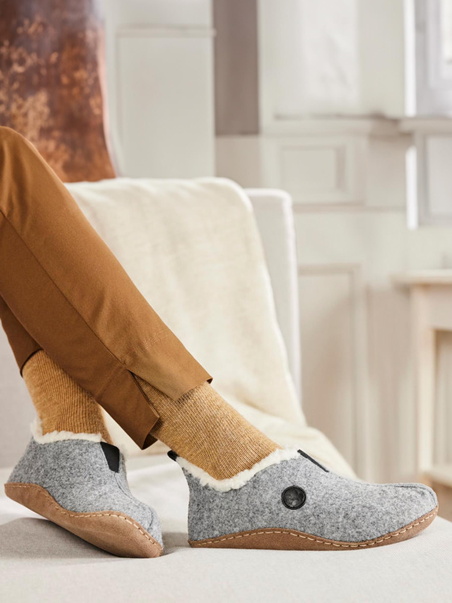 En person iført lysebrune bukser, lysebrune sokker og grå Rieker 04399 Cosy Innesko med kremfarget fôr slapper av i en lys sofa og nyter antistress-komforten fra Rieker-merket i et koselig innemiljø.