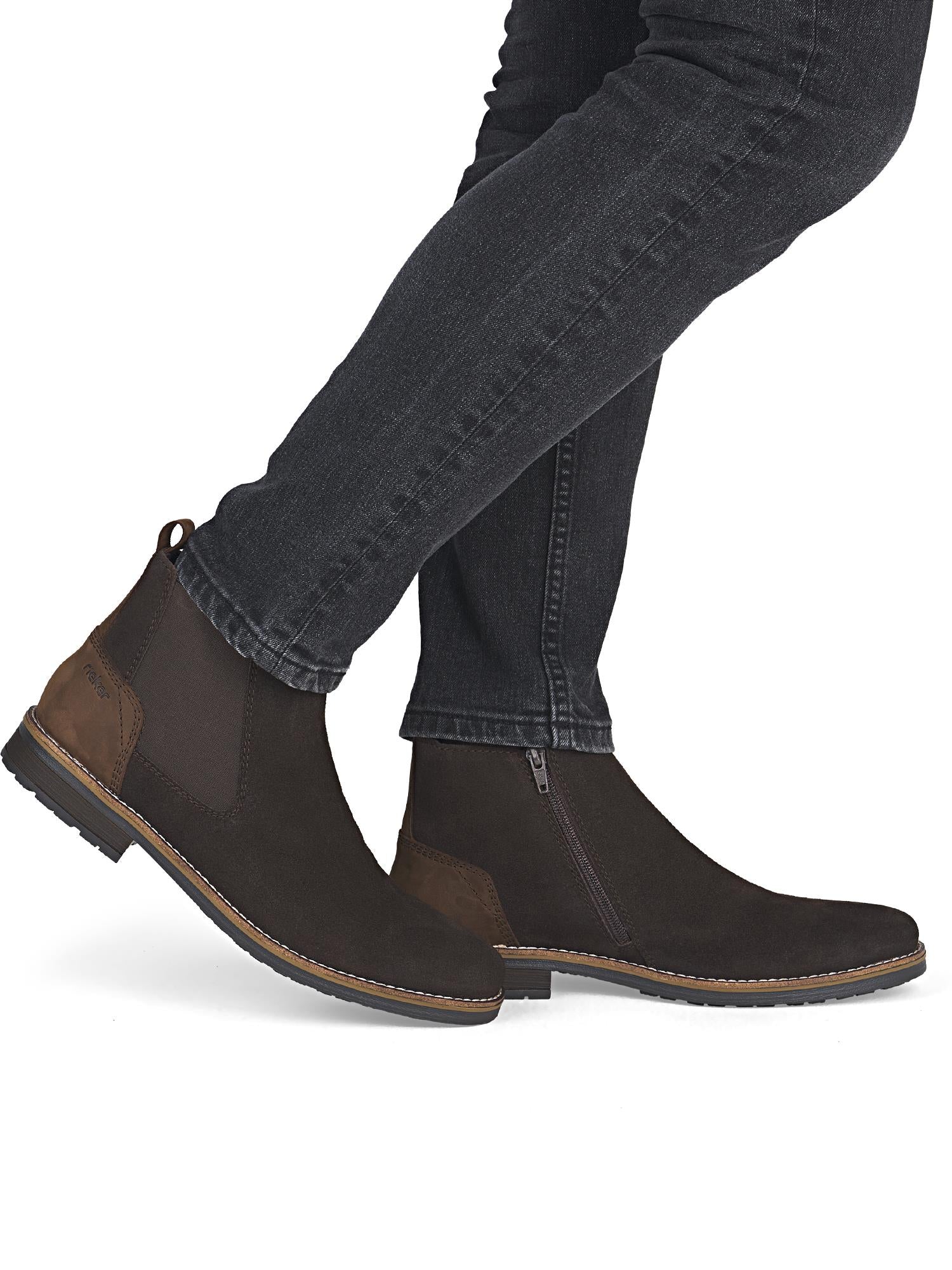 Personen har på seg 04245 Rieker Donovan lærstøvler fra Rieker Comfort & Style, med glidelåser på siden og elastiske paneler, sammen med sorte jeans på en hvit bakgrunn.