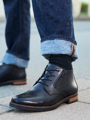 Et nærbilde viser en person i mørkeblå jeans med mansjett, svarte sokker og Rieker Comfort & Style 04231 Mantova Boot Black med ekstra bred passform, som står på et fortau.