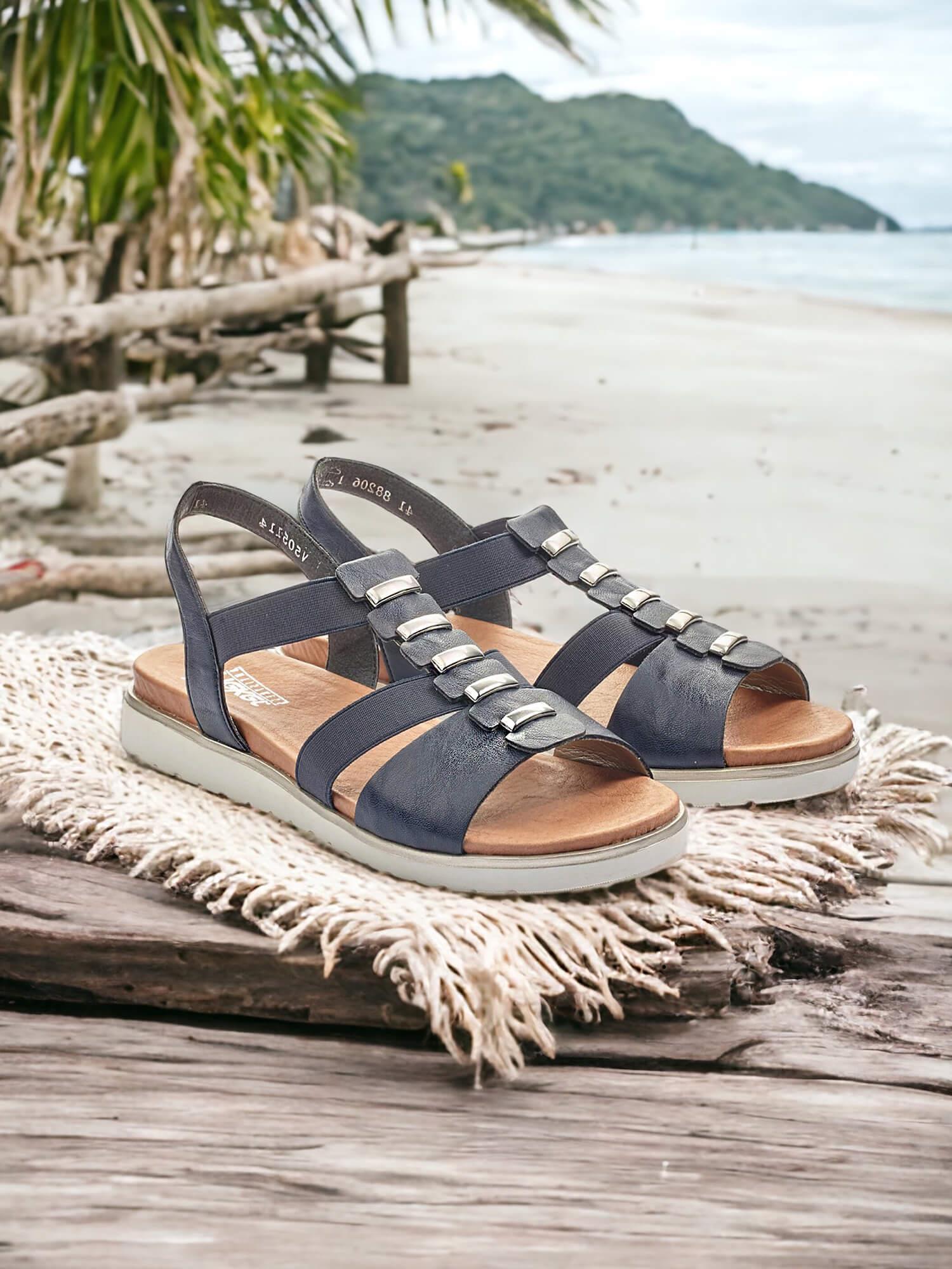 04016 Rieker Detailed Elastic Sandal fra Rieker Comfort & Style, i svart syntetisk skinn med spenner, hviler på en vevd matte over drivved på en sandstrand med palmer og åser i bakgrunnen.