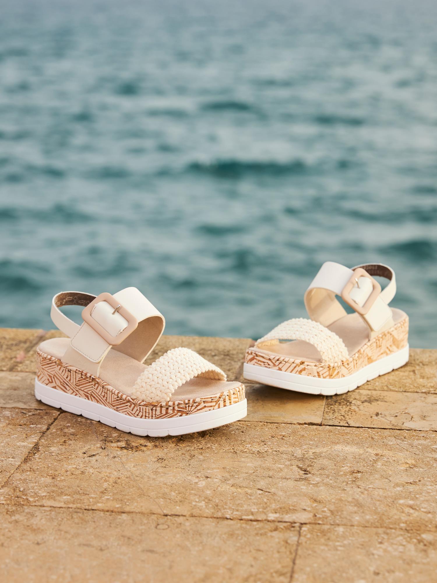 Et par beige 08091 Rieker Tropic-sandaler fra Rieker Comfort & Style, med tykke hvite såler, vevde detaljer og antistress-teknologi for ekstra komfort, hviler på en steinoverflate nær havet.