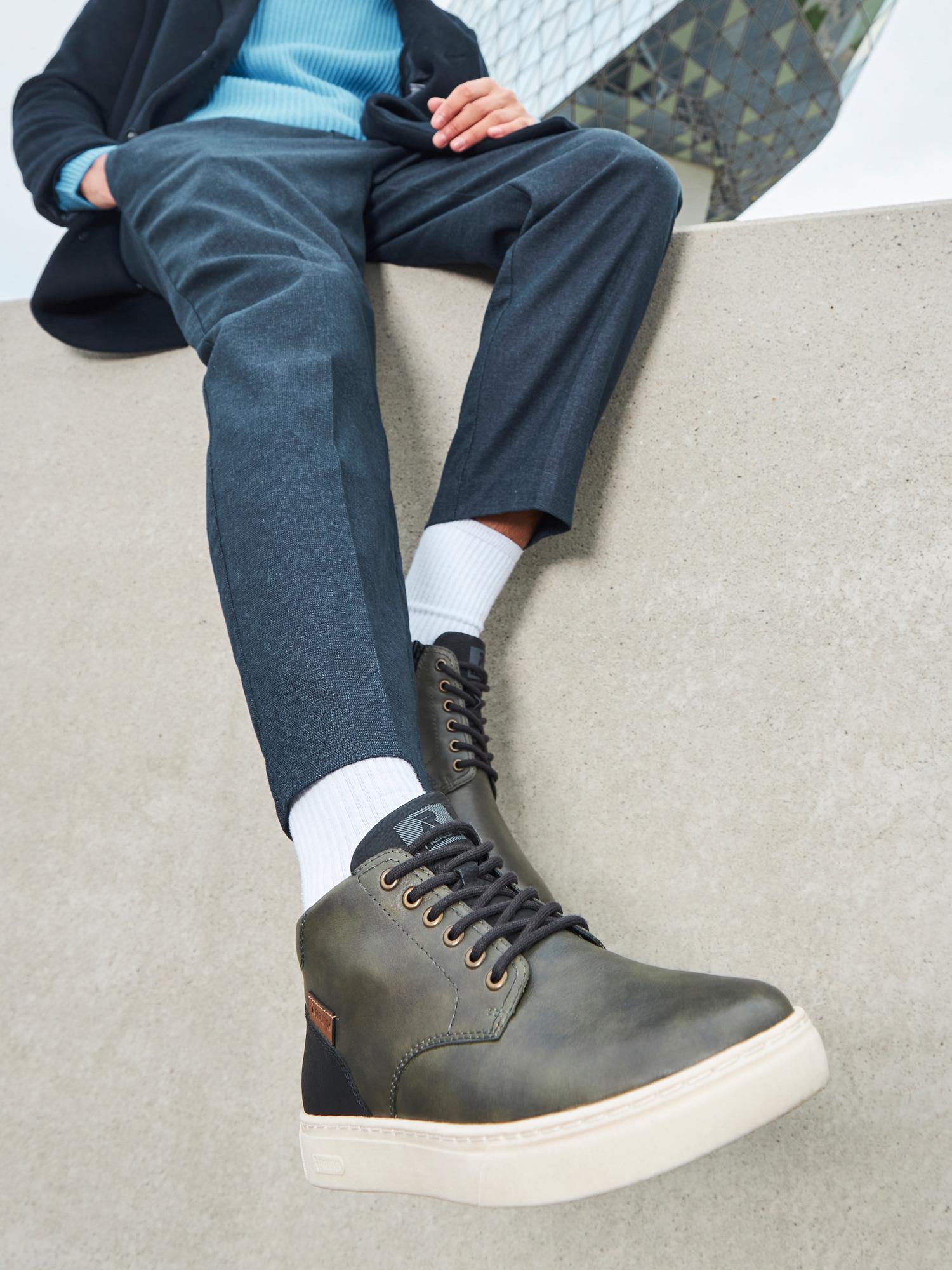 En person iført blå bukse, svart frakk, hvite sokker og Rieker-Evolution 04518 Forest Green Leather RevolutionSoft joggesko med hvit såle sitter på en betongkant med utstrakte ben foran en moderne bygning.