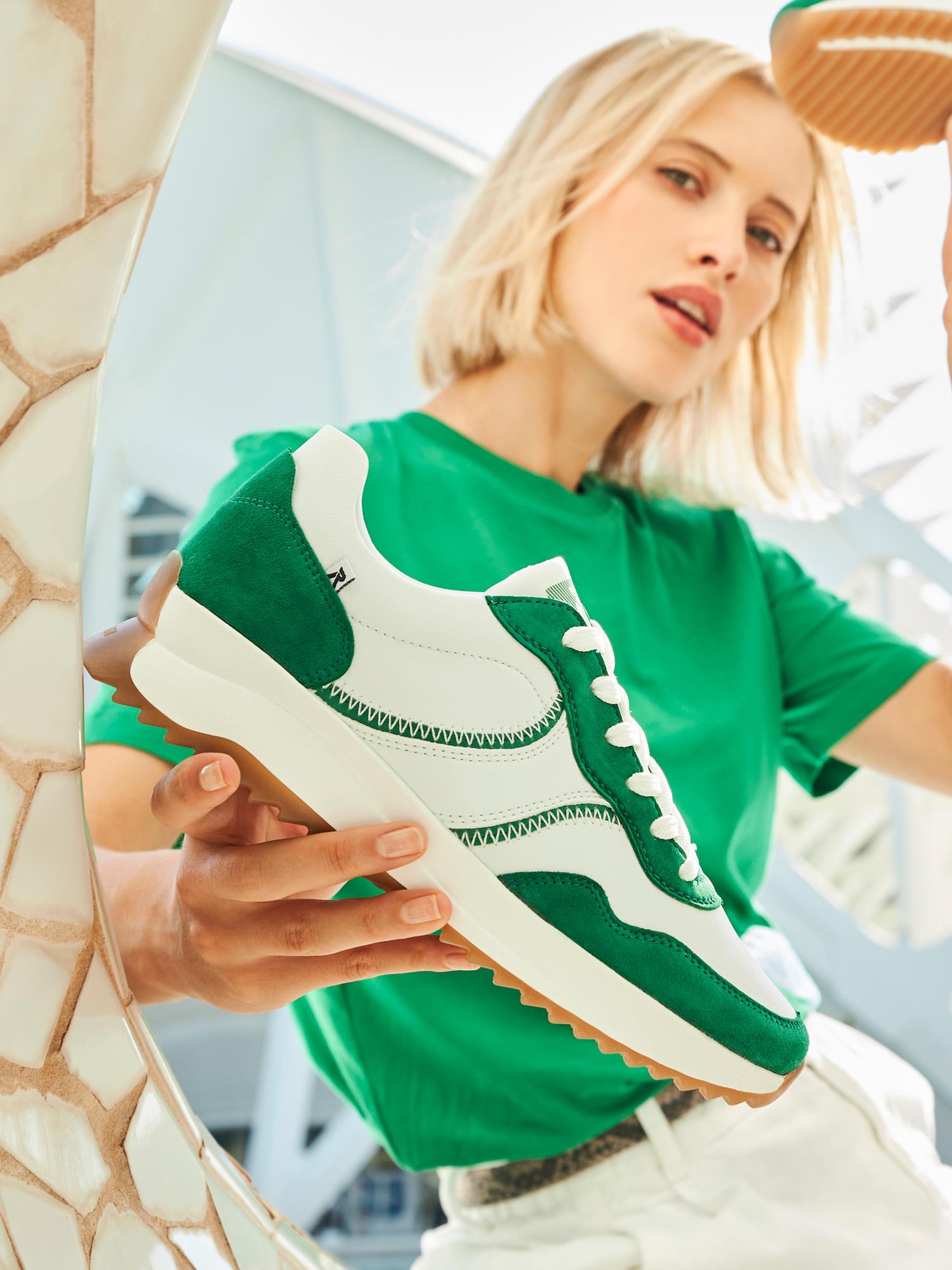 En kvinne i en grønn skjorte holder 07634 Rieker-Evolution Emerald Leather Sneaker fra Rieker Sport mot kameraet. Det glatte skinnet og den komfortable vidden blander moderne stil og brukervennlighet. Samtidsarkitekturen er uskarp i bakgrunnen.