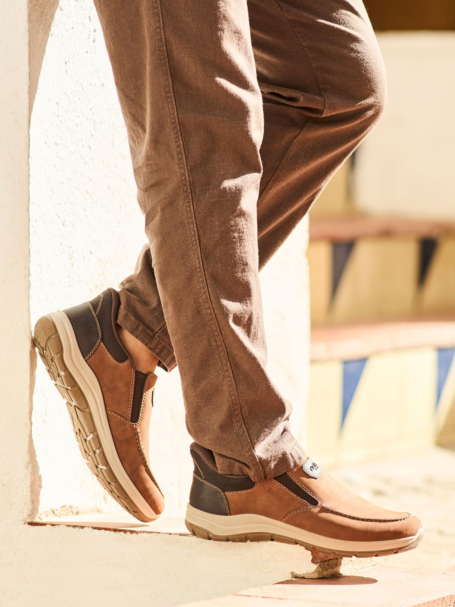 En person iført brune bukser står med den ene foden mod en hvit væg i sollys iført 02862 Rieker FlexStep Slip-Ins fra Rieker Comfort & Style med beige såler, antistress-teknologi og mørkebrune detaljer.