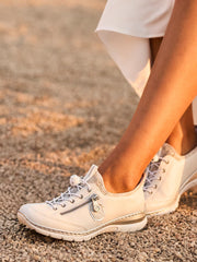 En person iført 03150 Rieker EasyMove Slip-on White joggesko fra Rieker Comfort & Style, med IM-EVA-såle og glidelåsdetaljer, står på grus med deler av det hvite antrekket sitt synlig.
