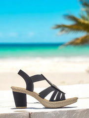 07498 Rieker Feather Heeled Sandal fra Rieker Comfort & Style vises utendørs med en vevd såle og svart stroppedesign, plassert mot hvit sand, turkis hav, palmeblader og en knallblå himmel – som forener stil og komfort.