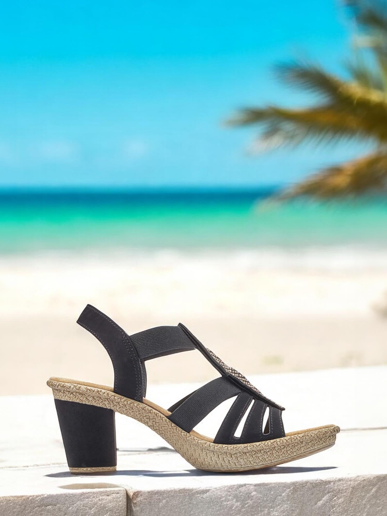 07498 Rieker Feather Heeled Sandal fra Rieker Comfort & Style vises utendørs med en vevd såle og svart stroppedesign, plassert mot hvit sand, turkis hav, palmeblader og en knallblå himmel – som forener stil og komfort.