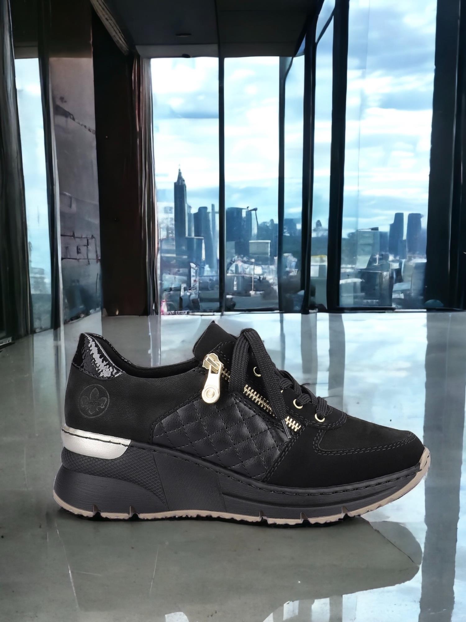 Et par 04389 Rieker Schwarz Kilesneakers fra Rieker Comfort & Style, med vatterte sider og glidelås, er utstilt innendørs på et skinnende gulv med store glassvinduer som avslører byens skyline.