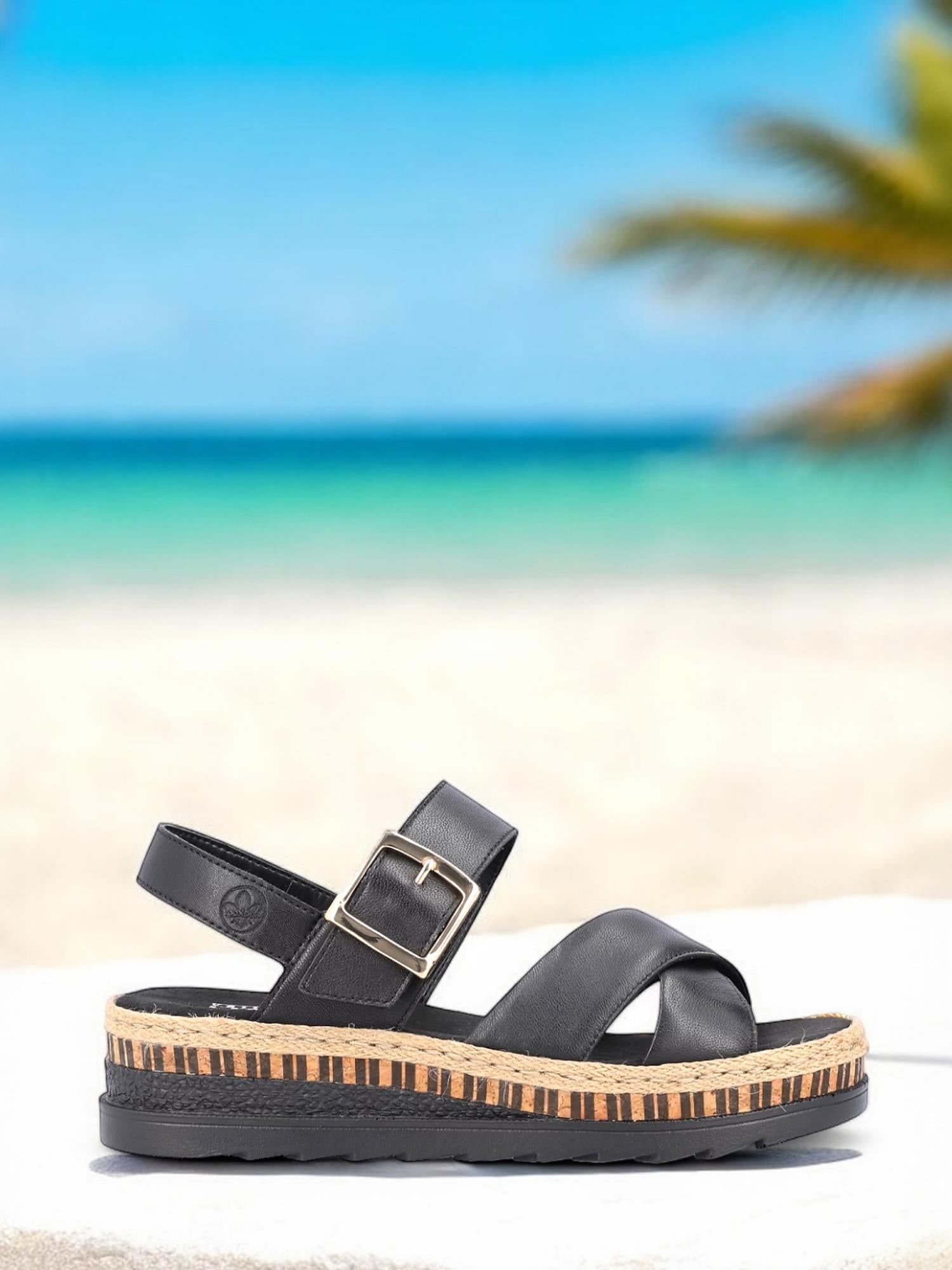 Rieker Comfort & Style 08084 Arizona Sandal i svart skinn, med optimal komfort og en plateausåle med korkstriper, hviler på en hvit overflate med en uskarp strand, turkis hav og palmer under en klarblå himmel.
