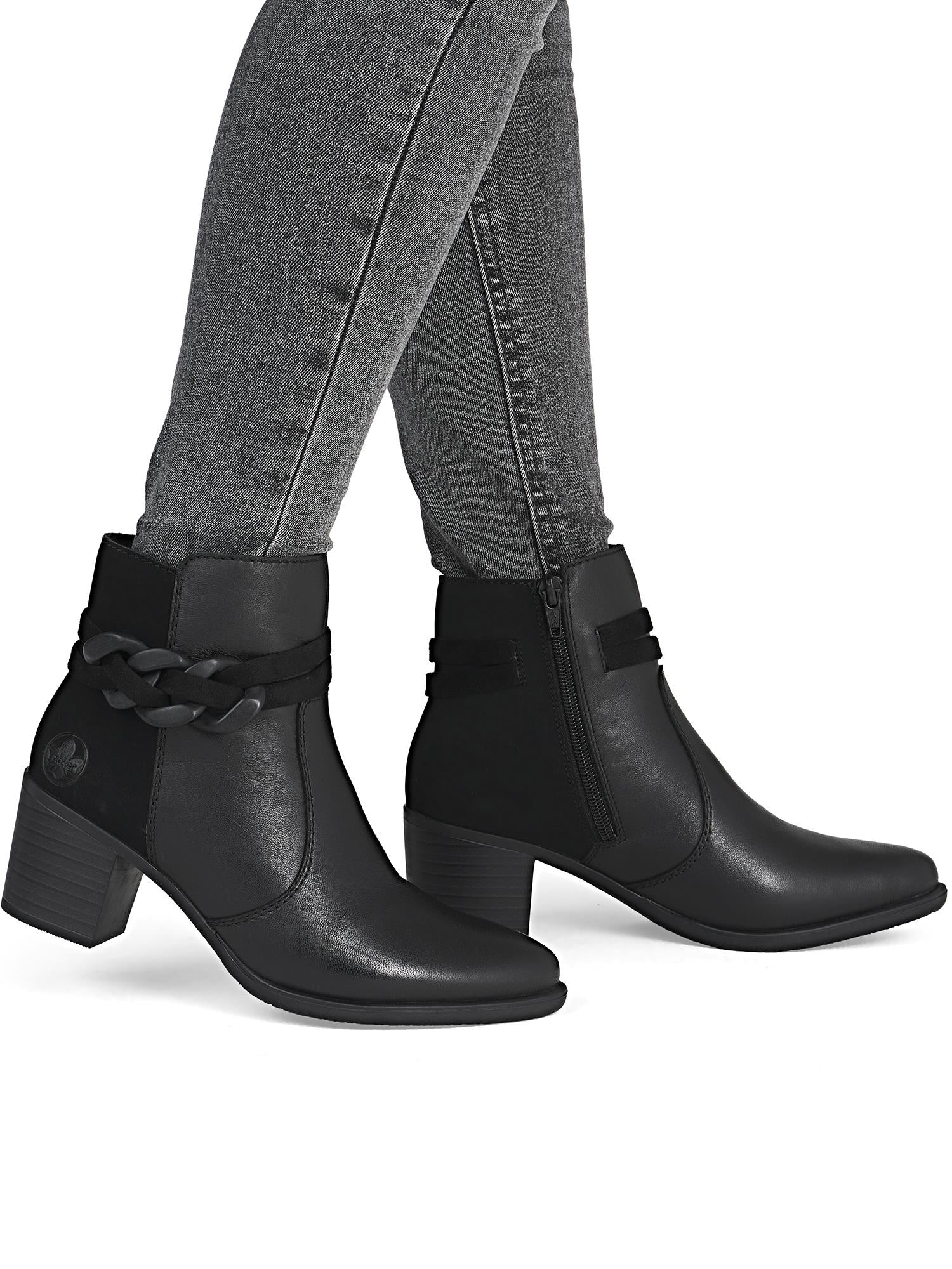 En person i grå skinny jeans og 05072 Rieker Amara Ankle Boot fra Rieker Comfort & Style - en elegant svart ankelstøvel med flettet stropp og antistress-teknologi - står mot en hvit bakgrunn.