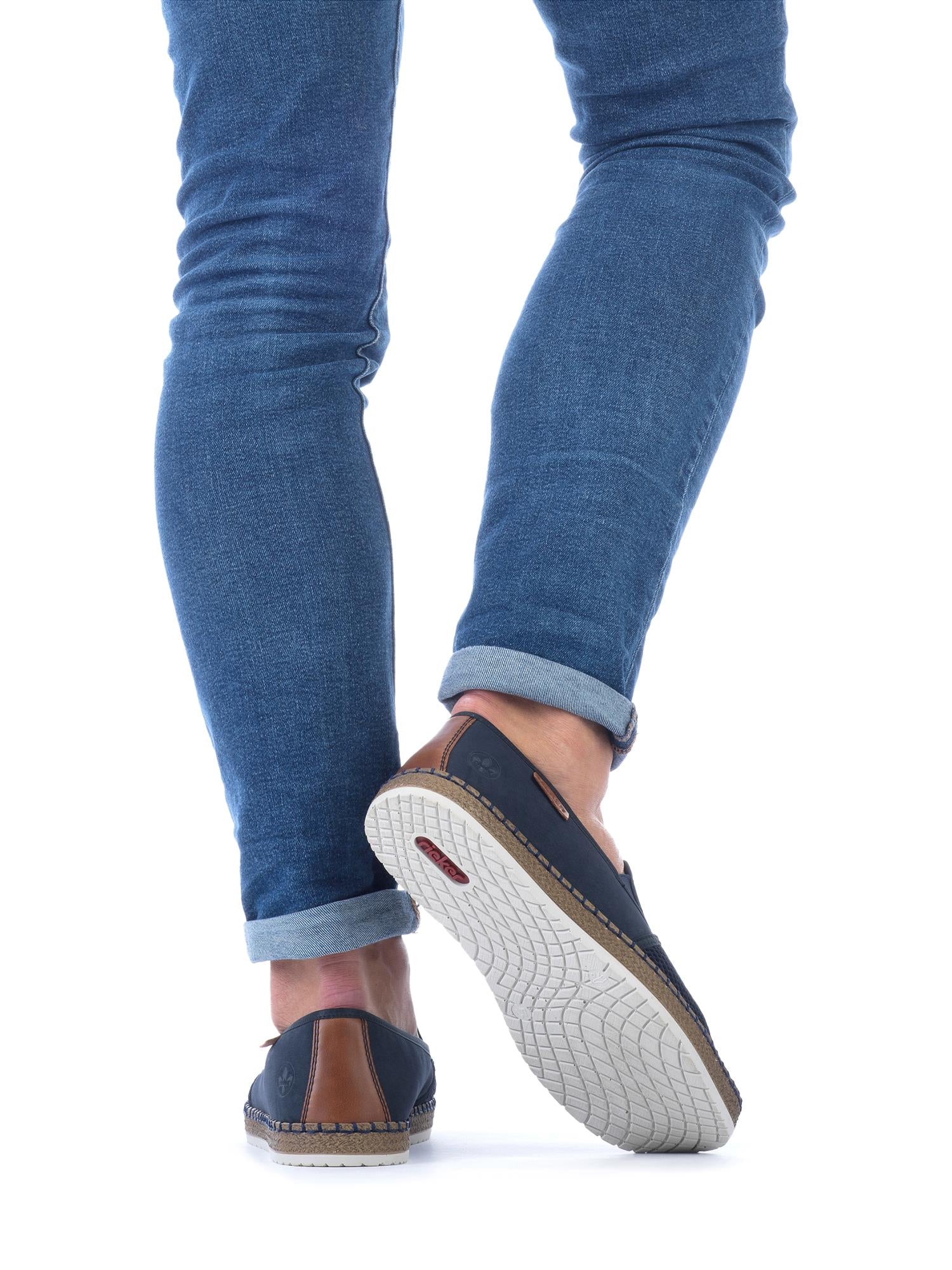 En person i blå jeans med mansjett og marineblå 07510 Rieker Coastline Moc slip-ons fra Rieker Comfort & Style går bort, vist bakfra på en hvit bakgrunn.