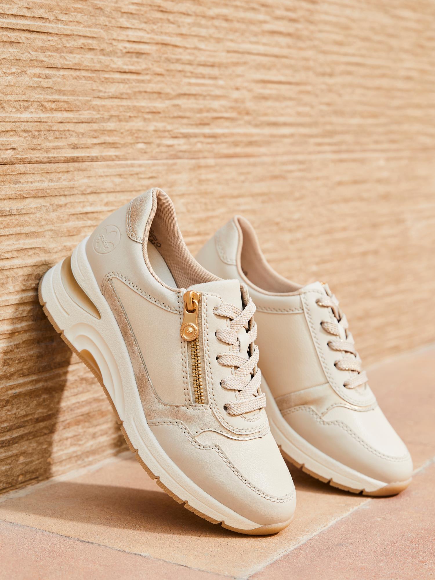 Et par 07583 Rieker Reflect Leather Zip Sneakers fra Rieker Comfort & Style i beige, med hvite såler, snøring foran, uttakbar innersåle og glidelåser på sidene, satt mot en strukturert brunfarget vegg og gulv.