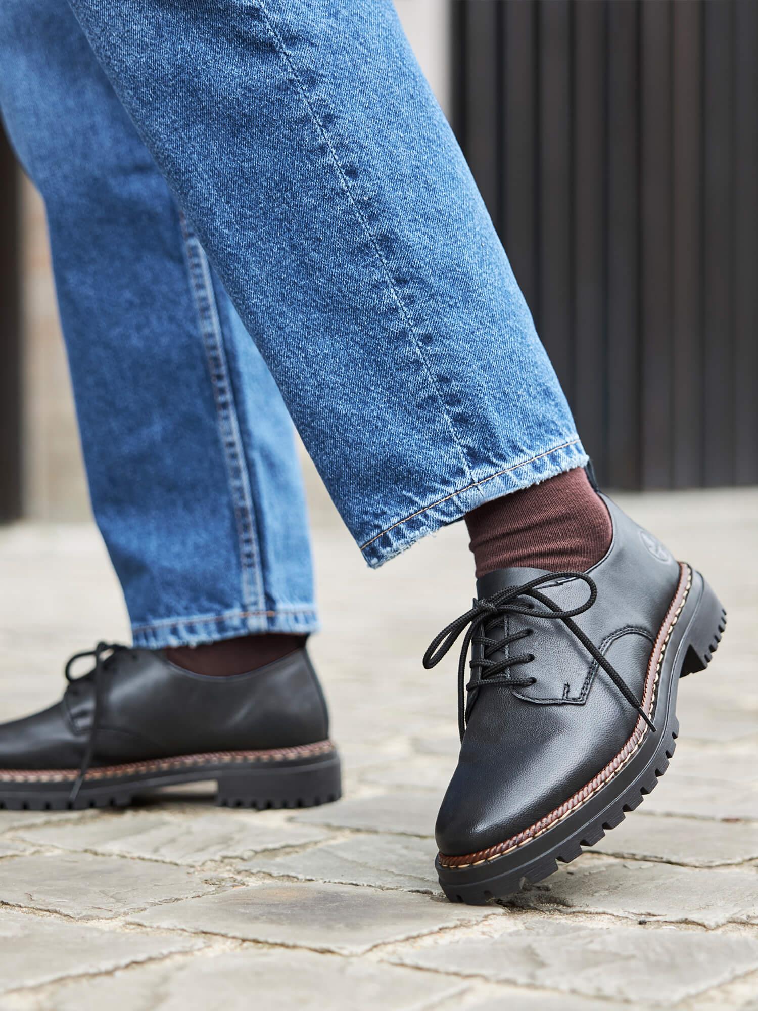 En person i blå jeans har på seg Rieker Comfort & Style 04377 Classic svarte sko med tykke, robuste såler og skinnende overdel, står på et steinbelegg; bare underbena og føttene er synlige.