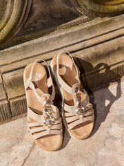 Et par beige 08090 Rieker Crystal Shell-sandaler fra Rieker Comfort & Style, med juvelutsmykninger og elastiske reimer, utstilt på en solbeskinnet stein nær en utsmykket vegg.