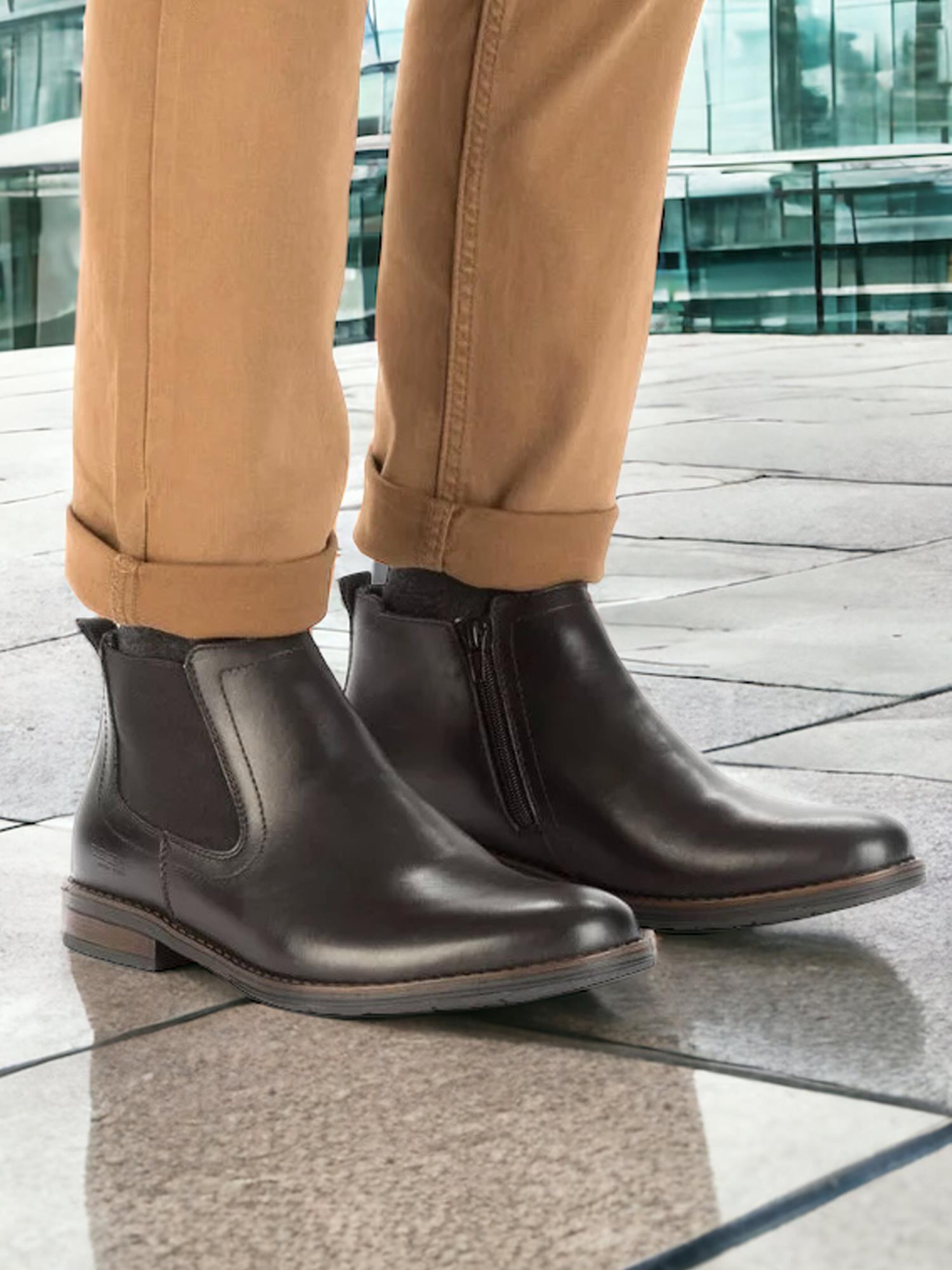 En person iført solbrune bukser og Rieker 04404 Black Short Boot Leather med antistresssåle står på en flislagt uteplass, med en moderne glassbygning i bakgrunnen.