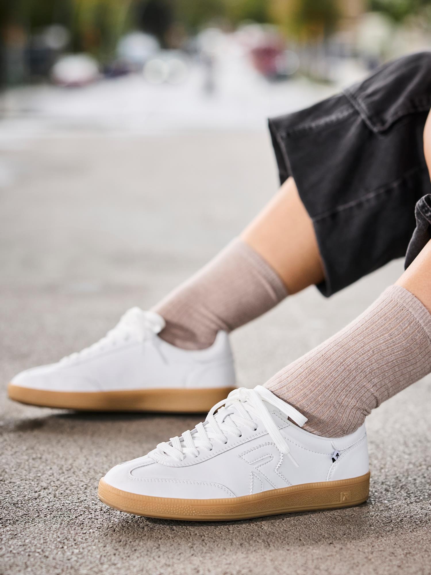 En person med lysebrune sokker og korte, svarte bukser sitter på et fortau, iført hvite Rieker Sport 03486 San Diego Leather Sneakers med gummisåler, et påsydd R-design og Swing2Go-teknologi. Bakgrunnen er uskarp.
