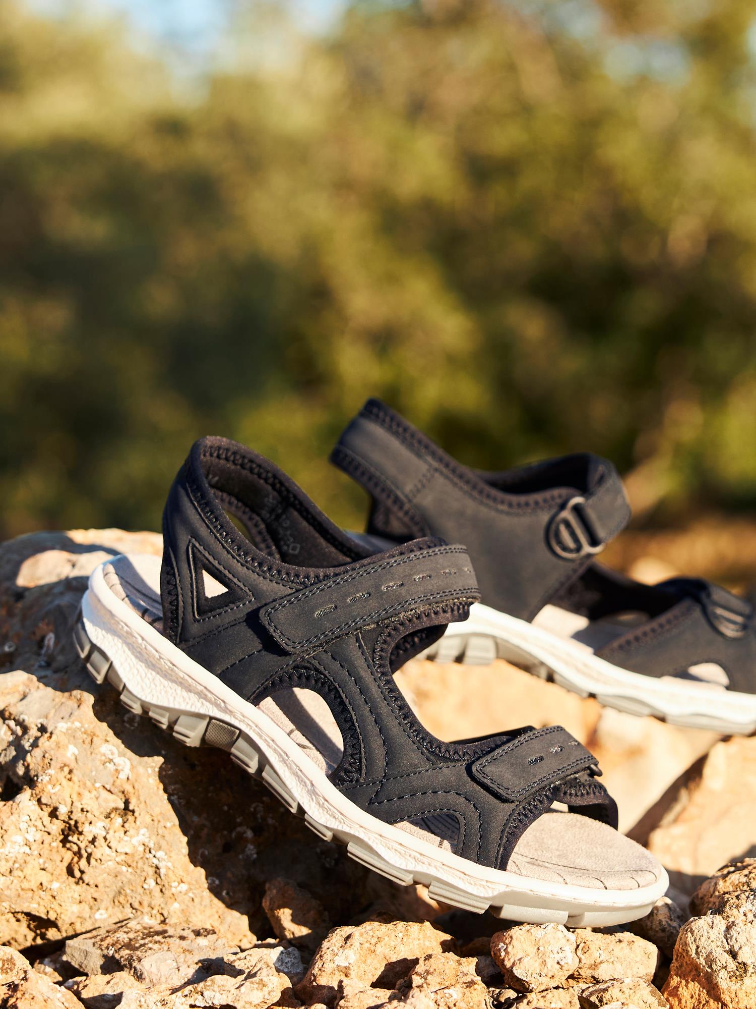 03009 Rieker Marine ComfortWave Sandal fra Rieker Comfort & Style har borrelåsstropper og hviler på solfylte steiner, noe som lover komfort hele dagen. Uklart grønt løvverk i bakgrunnen gir et snev av ro.