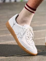 En person i Rieker 03336 San Diego Leather Sneaker Herre Hvit står på et steinbelagt fortau, iført beige ribbede sokker med to røde striper - klar for behagelige skritt i klassisk Rieker-stil med gummisåler.