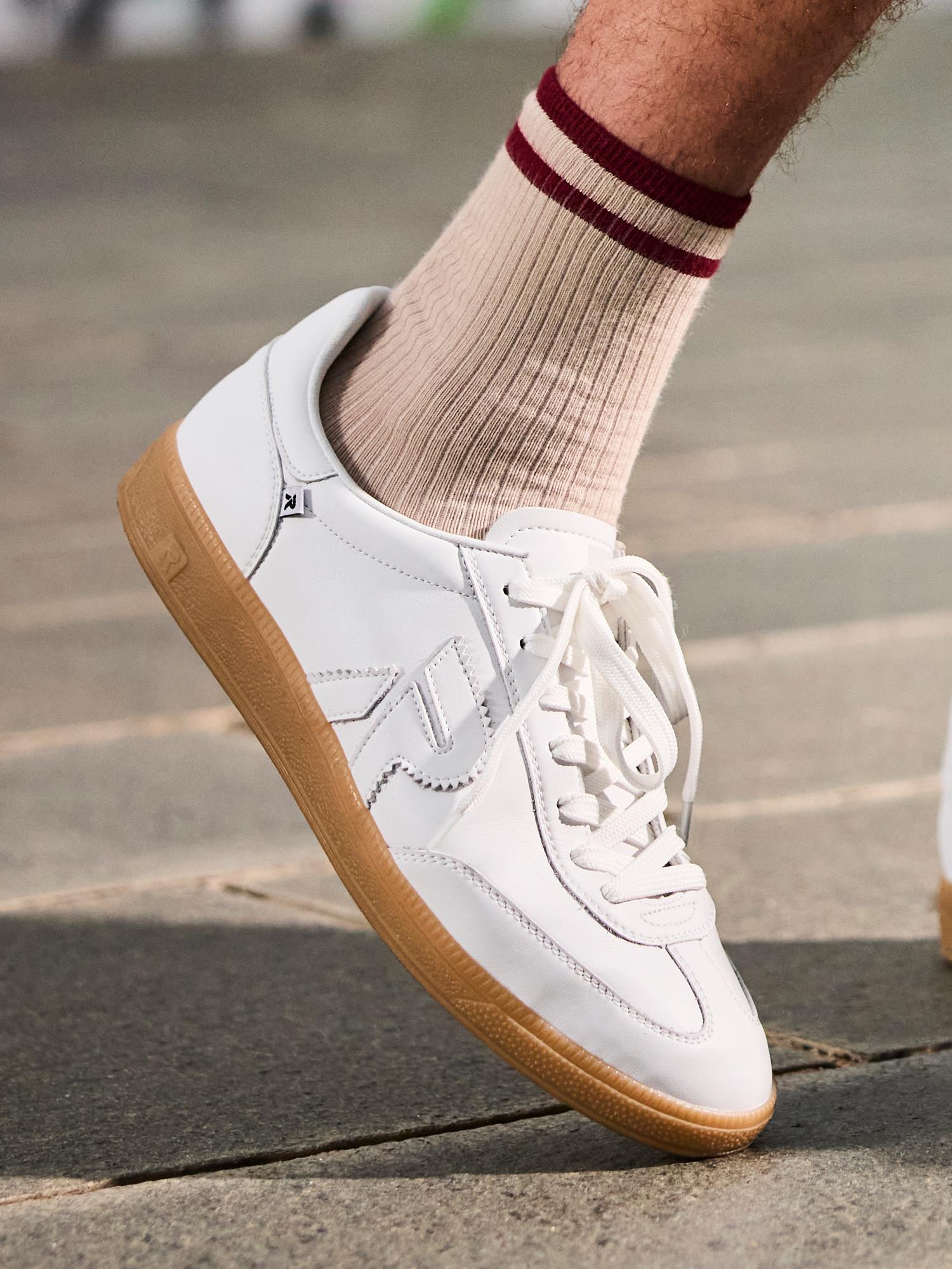 En person i Rieker 03336 San Diego Leather Sneaker Herre Hvit står på et steinbelagt fortau, iført beige ribbede sokker med to røde striper - klar for behagelige skritt i klassisk Rieker-stil med gummisåler.
