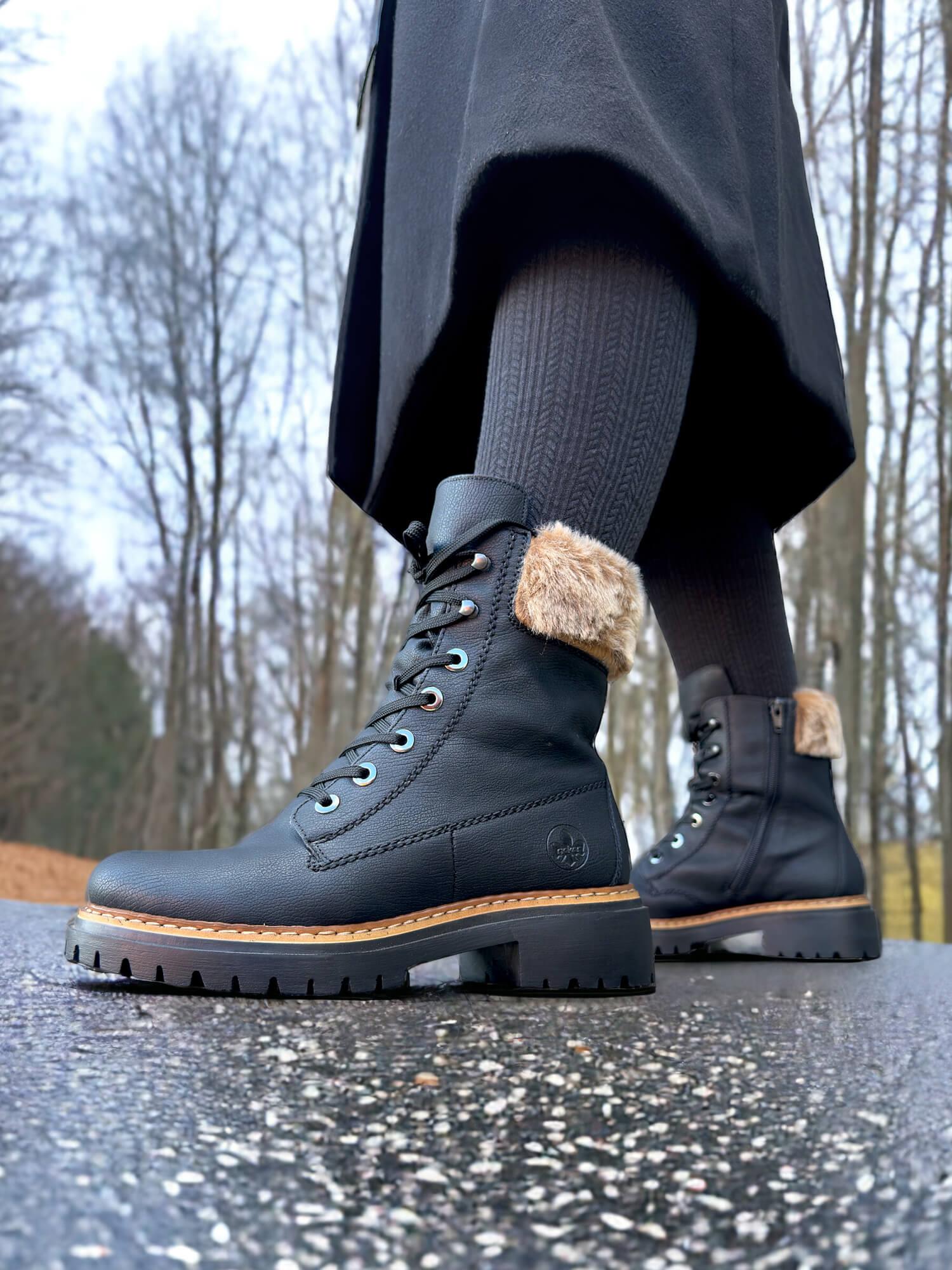 Et nærbilde av en person iført svarte 05377 Rieker pelsstøvler fra Rieker Comfort & Style, stående på vått fortau. Bare trær og en overskyet himmel er uskarpt i bakgrunnen.