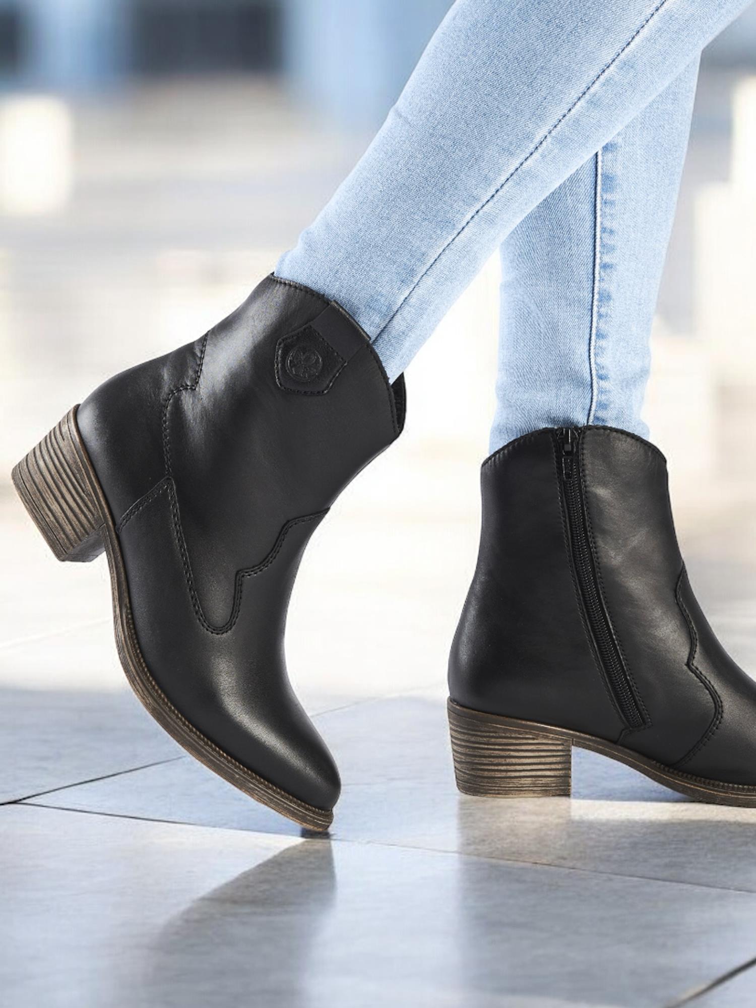 En person iført lyseblå skinny jeans og 04859 Rieker Leather Cowgirl Boots fra Rieker Comfort & Style, med lave, høye hæler og glidelåser på siden, står på et flislagt gulv.