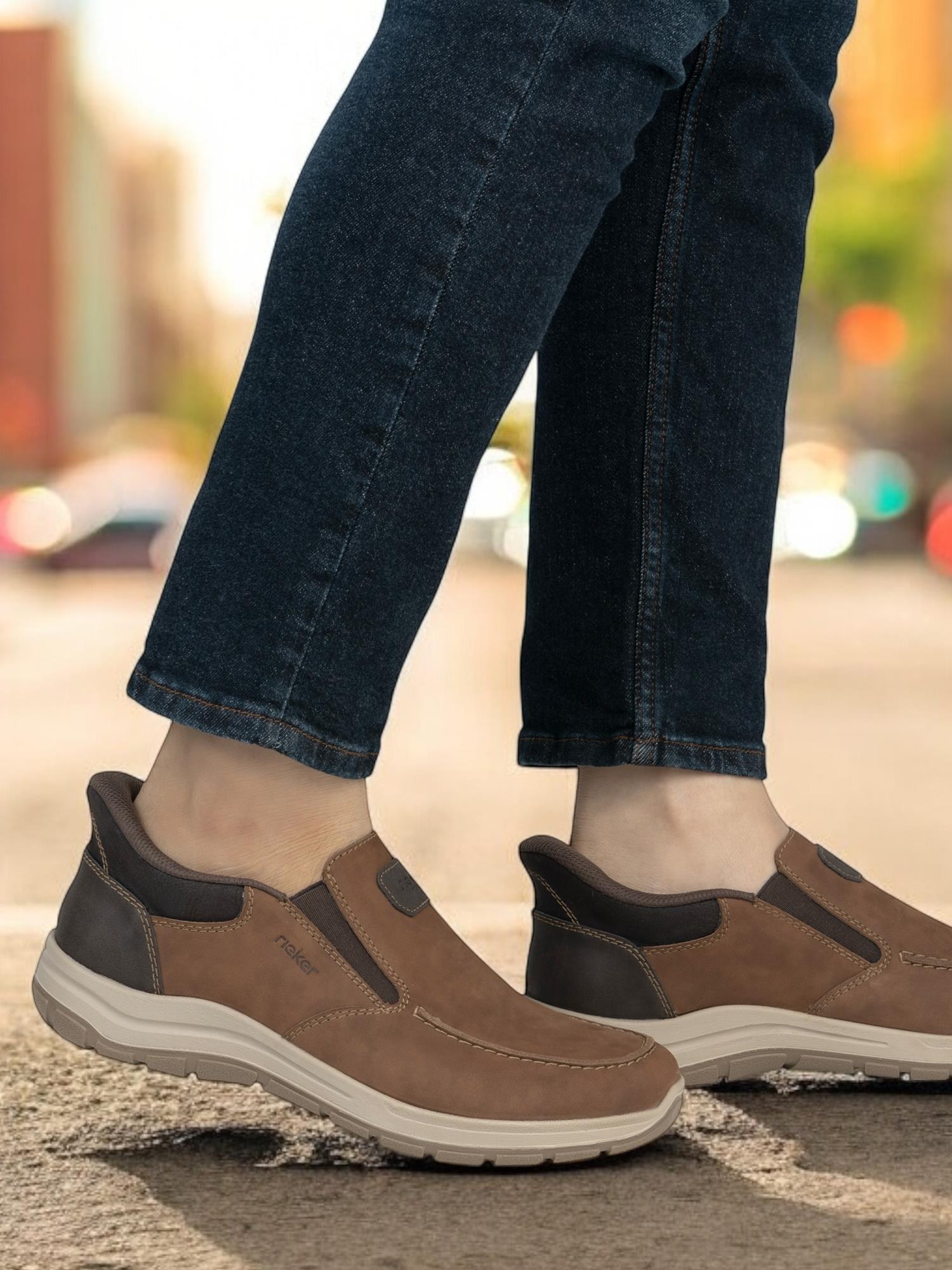 På en bygate nyter noen i mørkeblå jeans og brune 02862 Rieker FlexStep Slip-Ins fra Rieker Comfort & Style antistress-teknologisk komfort blant uskarpe bygninger og grøntområder.
