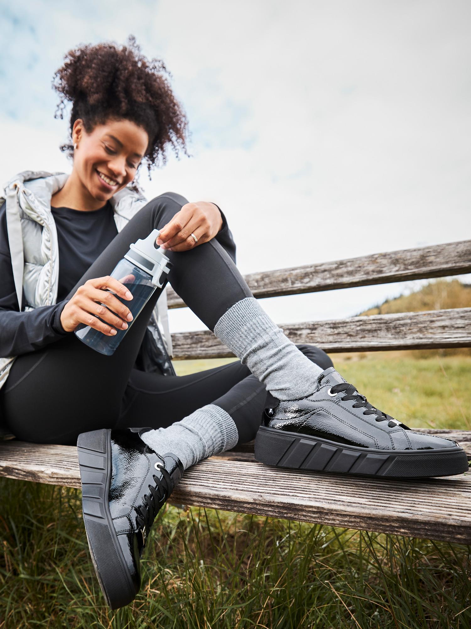 En smilende kvinne i sportsklær og grå sokker sitter på en trebenk utendørs, med beina utstrakt for å vise frem de svarte 04543 Rieker-Evolution Patent Leather Platform Sneakers fra Rieker Sport. Gress og himmel fyller bakgrunnen.