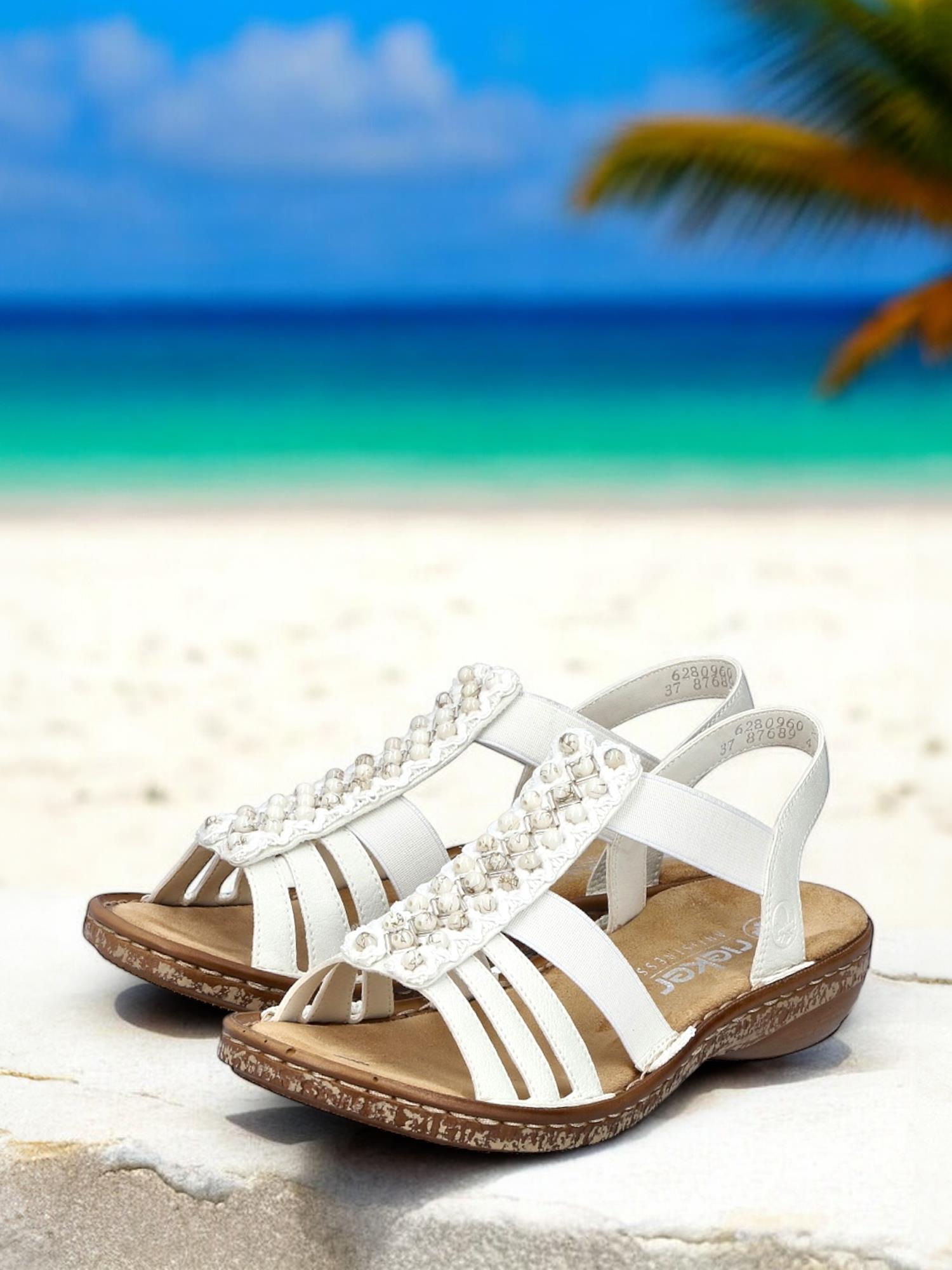 07496 Rieker Pearl White-sandalen fra Rieker Comfort & Style, med perledetaljer og brune såler, hviler på stein med en sandstrand, turkis hav og palmer mykt uskarpt i bakgrunnen.